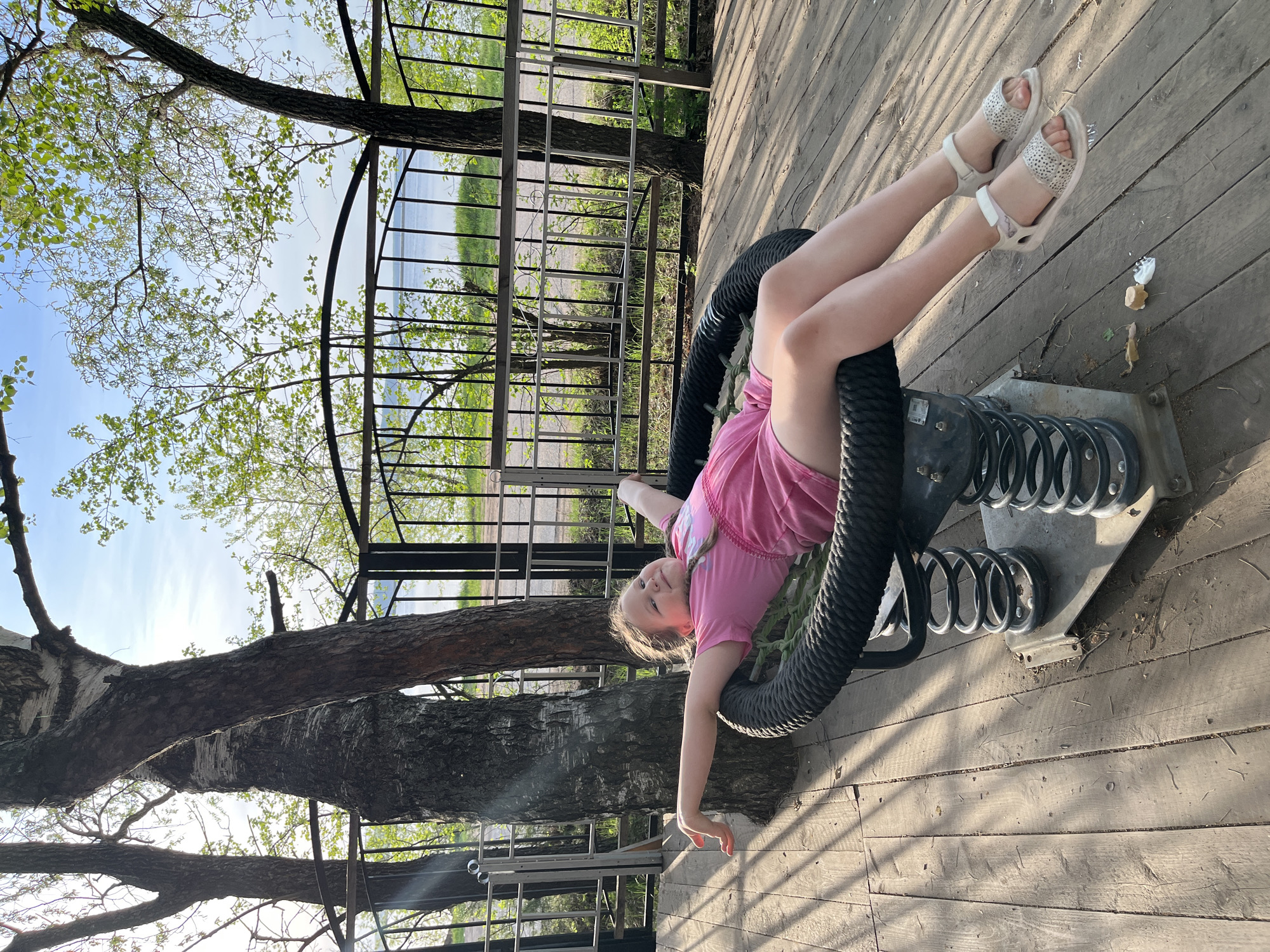 A young girl is on a black rope spring rider. The rider is on a wooden deck, with a metal railing in the background. There are trees in the background and to the side. The location is in the city of Chaikovsky, Russia. The girl is wearing a pink shirt and white sandals. She is holding onto the rope rider with her hands. The sun is shining and casting shadows on the deck.