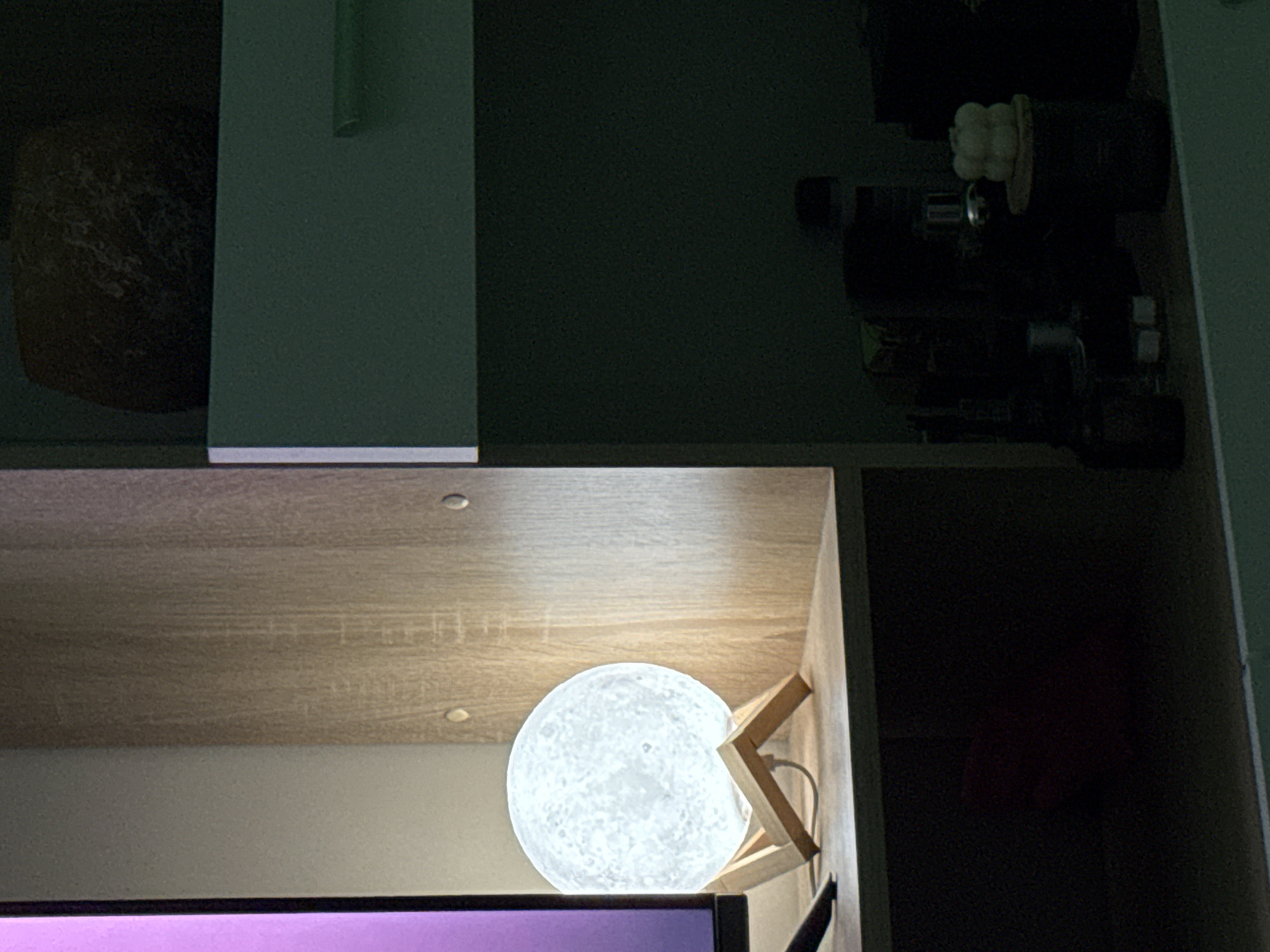 The image displays an indoor scene, likely at night, focusing on a glowing spherical moon lamp placed on a wooden shelf. The lamp, characterized by its textured, crater-like surface, rests on a light-colored wooden stand within a cubby of a shelving unit. It emits a bright, white light that illuminates the light brown wooden shelf above it.
Below the shelf holding the moon lamp, the top edge of a monitor screen is visible, emitting a soft purple light. The shelving unit itself is constructed from light brown wood with a noticeable grain. Above the moon lamp's compartment, another section of the shelving unit is visible but largely in shadow. On the far left of this upper section, a dark, textured, possibly round object is faintly discernible. To the right, several dark, indistinct items, potentially bottles or small electronics, are stacked. A small, white, ribbed object, possibly a decorative piece or candle, is visible among these dark items. A dark green or grey vertical panel or wall is prominent on the upper left side of the frame. No people or text are visible in the image.