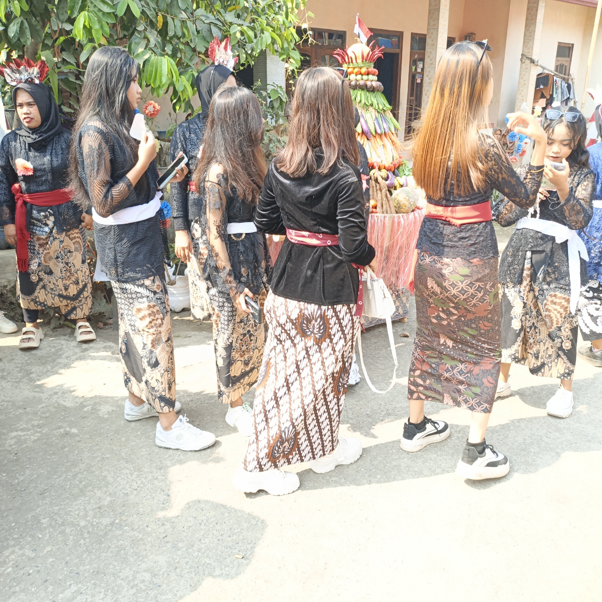 In the city of Tanahbaya, Indonesia, a group of young women in traditional attire are gathered outdoors. Their clothing consists of black lace tops and batik skirts, with some wearing red or white sashes. The women are diverse in their engagement with the scene; some are looking towards a decorative display of fruits and vegetables arranged in a conical shape, while others are holding cell phones. One woman is adjusting her sunglasses, and another is holding a tissue. The environment appears to be a street or courtyard, with a building visible in the background and lush green foliage overhead. The lighting suggests it is daytime, with the sun casting shadows on the concrete ground. The overall atmosphere seems to be one of a local event or festival.