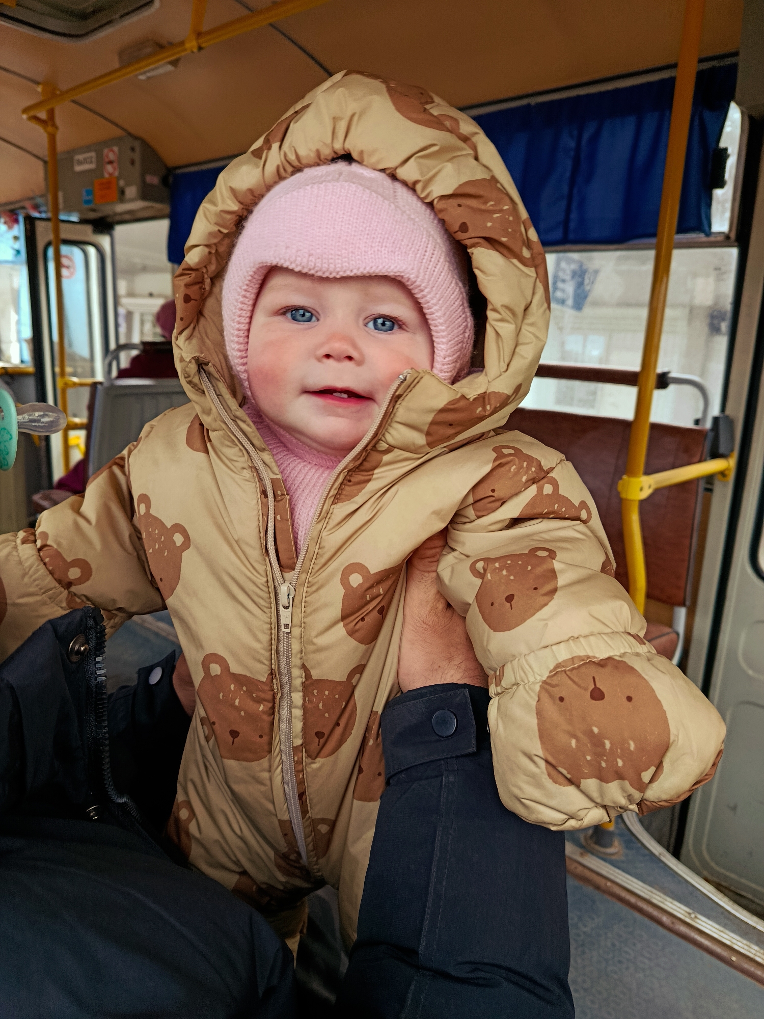 A young child wearing a pink hat and a brown teddy bear print snowsuit stands on what appears to be public transportation. The child has blue eyes, a slight smile, and rosy cheeks. The person holding the child is wearing a dark-colored jacket. The background shows the interior of a bus or train with seats, handrails, and windows. The location is in a city of Unknown, Unknown.