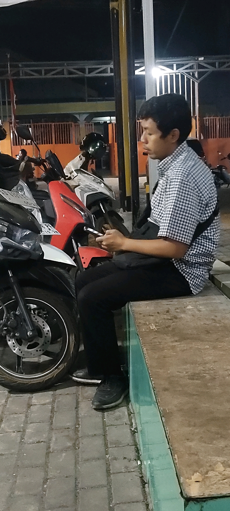 A young man with dark, short hair is seated on a wooden bench outdoors at night. He is wearing a black and white checkered collared shirt and black pants, with a black strap across his chest. He is looking down and holding a mobile phone in his right hand. He is positioned next to a red and white motorcycle. The bench is slightly elevated and appears weathered. The ground is paved with interlocking gray bricks. In the background, there are other motorcycles parked and an orange fence. The overall environment suggests an urban setting, potentially a parking area or a public space. The location is identified as Sungai Sipai, Indonesia.