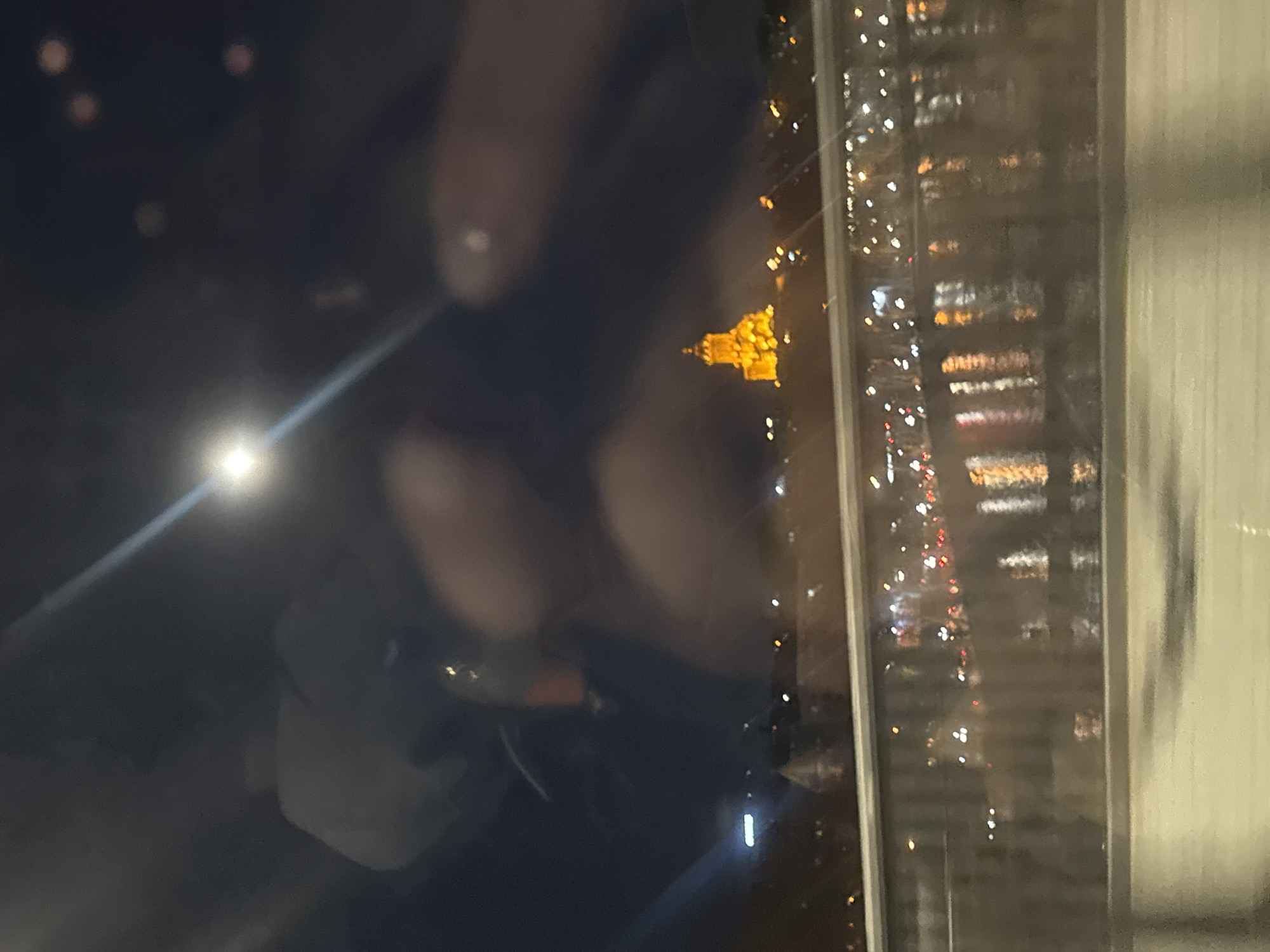 The image captures a nighttime cityscape viewed through a window, significantly influenced by reflections from the interior. The primary orientation appears to be landscape, though the provided image is rotated 90 degrees clockwise. Describing it in its implied landscape orientation:
On the left side of the frame, prominent reflections are visible on the window glass. A bright, circular light source, likely from a phone screen or an indoor lamp, is reflected in the bottom-left corner, emitting a strong lens flare that extends diagonally upwards and to the right across the reflection of the glass. Above and around this light, the blurred reflections of a hand and fingers are clearly discernible, with some fingers showing what appears to be light-colored nail polish. Other indistinct, darker shapes in this area suggest reflections of people or objects inside the room.
Through the translucent reflections, the exterior scene reveals a vibrant urban environment at night. Towards the center-right of the image, a distinctive building stands out, brightly illuminated in a golden-yellow hue. This structure features a prominent architectural design, possibly a dome or a multi-tiered roof, suggesting a significant landmark or historical building. To the right of this illuminated building and extending towards the far right edge of the frame, numerous small, sparkling lights indicate other buildings, possibly tall structures with many illuminated windows, and distant city lights, perhaps from streets or vehicles. A vertical window frame or mullion is visible on the far right, bordering the exterior view and separating it from what appears to be an interior wall or curtain on the extreme right.
The overall impression is a blend of interior and exterior, with the reflections creating a layered and somewhat abstract view. The time of day is clearly night, evidenced by the darkness and reliance on artificial lights. The perspective suggests a high vantage point, looking out over a city.