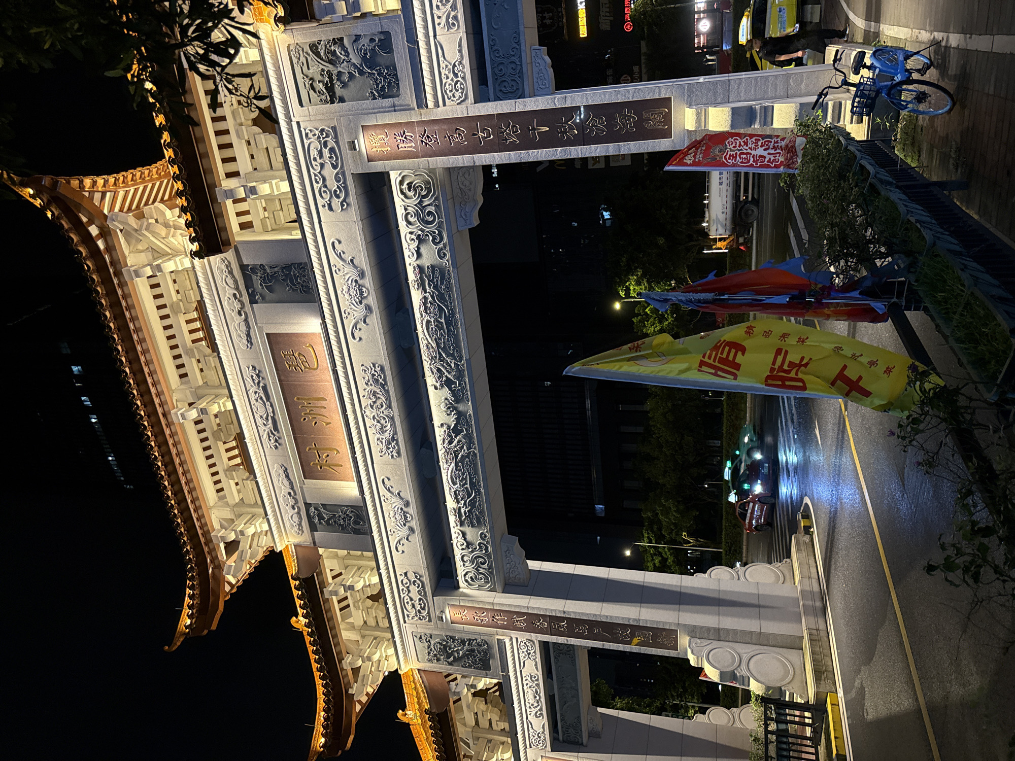 This is a night-time, eye-level shot of a traditional Chinese archway in Haizhu District, China. The archway is elaborately decorated with intricate carvings and illuminated by warm, artificial lights. Chinese characters are visible on plaques adorning the structure. In the foreground, there are several colorful flags planted in a planter box. To the right, a blue bicycle is parked on a paved walkway. Across the street, traffic lights indicate that vehicles are moving. A red car and a green car are visible in the street, with their headlights on. Beyond the archway, buildings are illuminated, suggesting an urban environment. The ground is wet, reflecting the lights, indicating recent rain. A person in dark clothing is standing near a yellow taxi on the right side of the image.