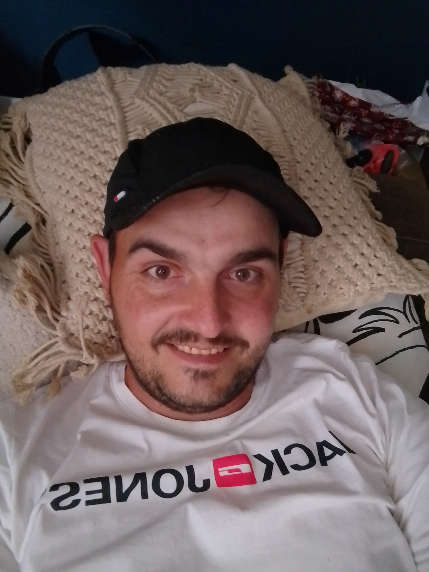 The media file is a selfie of a man lying down. He is wearing a white t-shirt with the words "JACK & JONES" printed on it in black lettering, with a pink rectangular logo next to the word "JONES." He is also wearing a black baseball cap. The man has short brown hair, brown eyes, and a short beard and mustache. He is smiling and looking at the camera.
The man is lying on a textured cream-colored blanket with fringe. Behind him, a dark blue object is visible, possibly a pillow or furniture. To the right of the man, there are other objects, including a red item and some patterned fabric, suggesting a casual, indoor setting. The lighting appears to be artificial or from an overcast sky, with no clear indication of time of day.
The overall emotion conveyed is relaxed and casual. The background elements suggest a domestic or personal space. There is no explicit information about the weather or specific location beyond "Wedel, Germany."