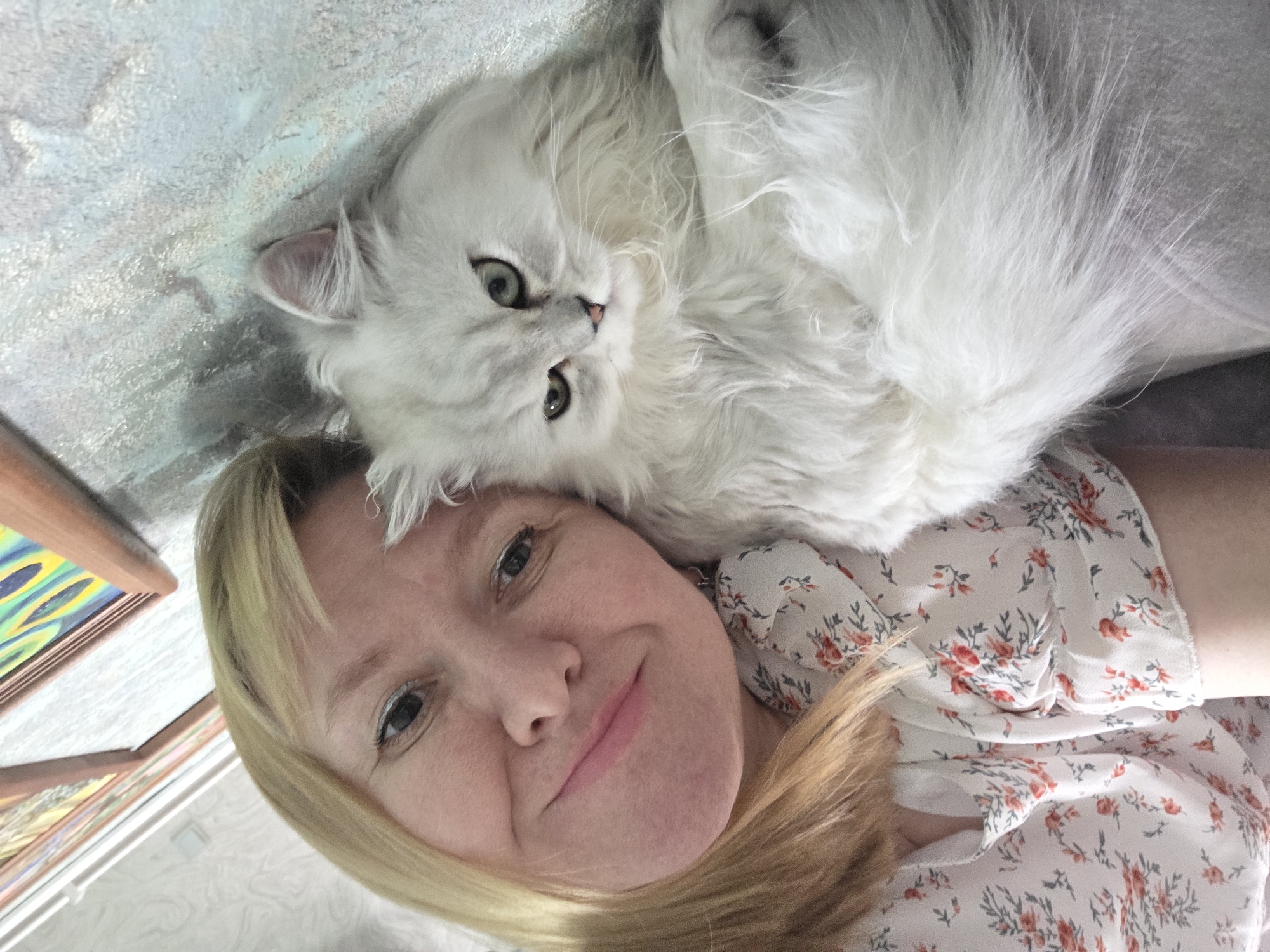 This is a selfie of a woman with blonde hair lying down with a fluffy white cat. The woman is looking up at the camera with a slight smile. The cat is lying on top of her, with its head resting on her shoulder and its body draped over her. The cat has green eyes and is looking at the camera. They are both on a light-colored, textured surface. In the background, there are framed pictures on the wall. The lighting suggests it is daytime. The scene appears to be indoors. The woman is wearing a light-colored dress with a floral pattern. The overall mood of the image is affectionate and relaxed.