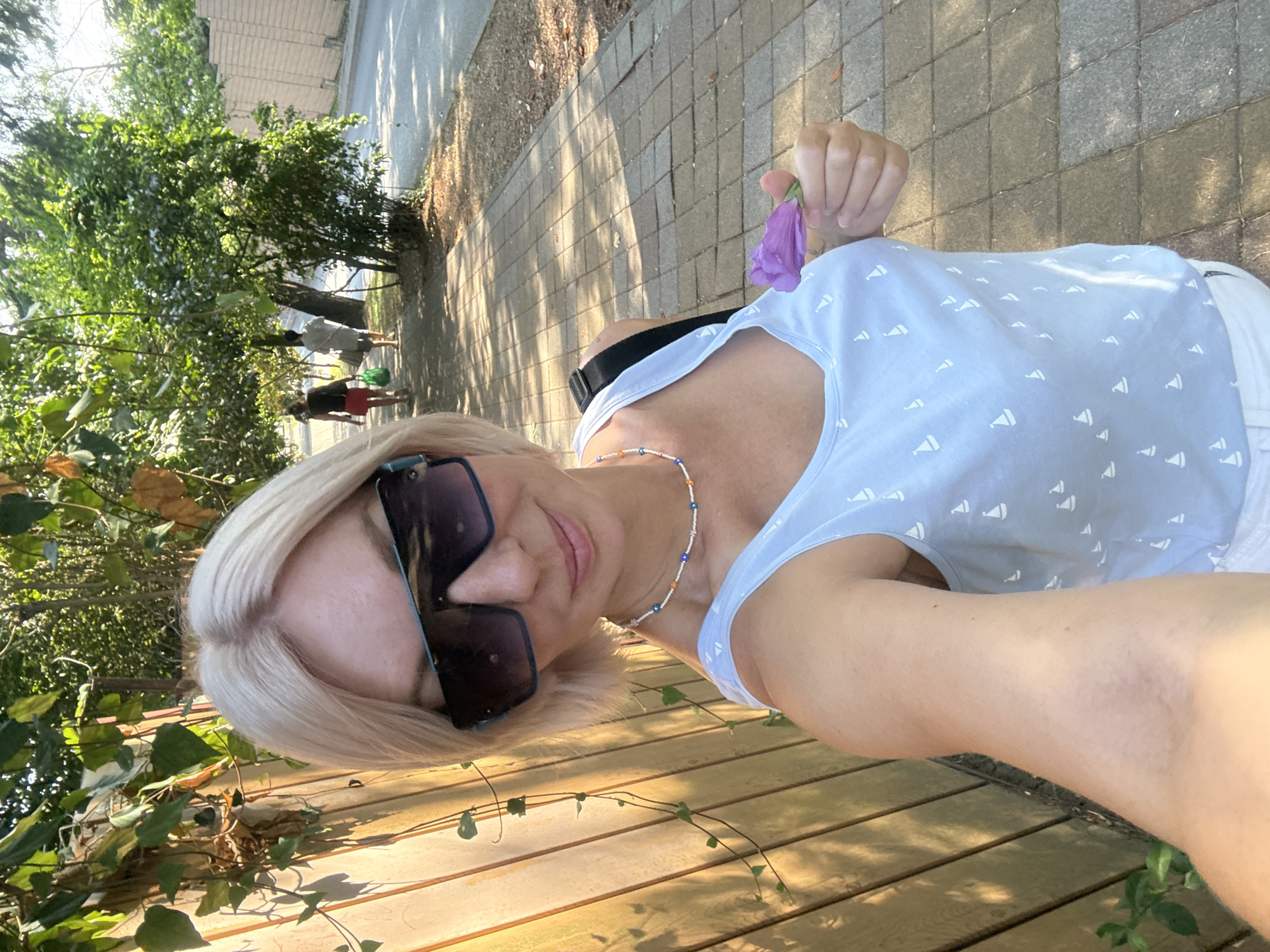 A woman with blonde hair and sunglasses takes a selfie outdoors. She is wearing a light blue tank top with a sailboat pattern and a beaded necklace. In her right hand, she holds a purple flower. Behind her, a paved sidewalk runs alongside a street lined with trees. In the background, other people are walking along the sidewalk. The scene appears to be in a park or a pedestrian area in Sochi, Russia, given the lush greenery and sunlit environment.
