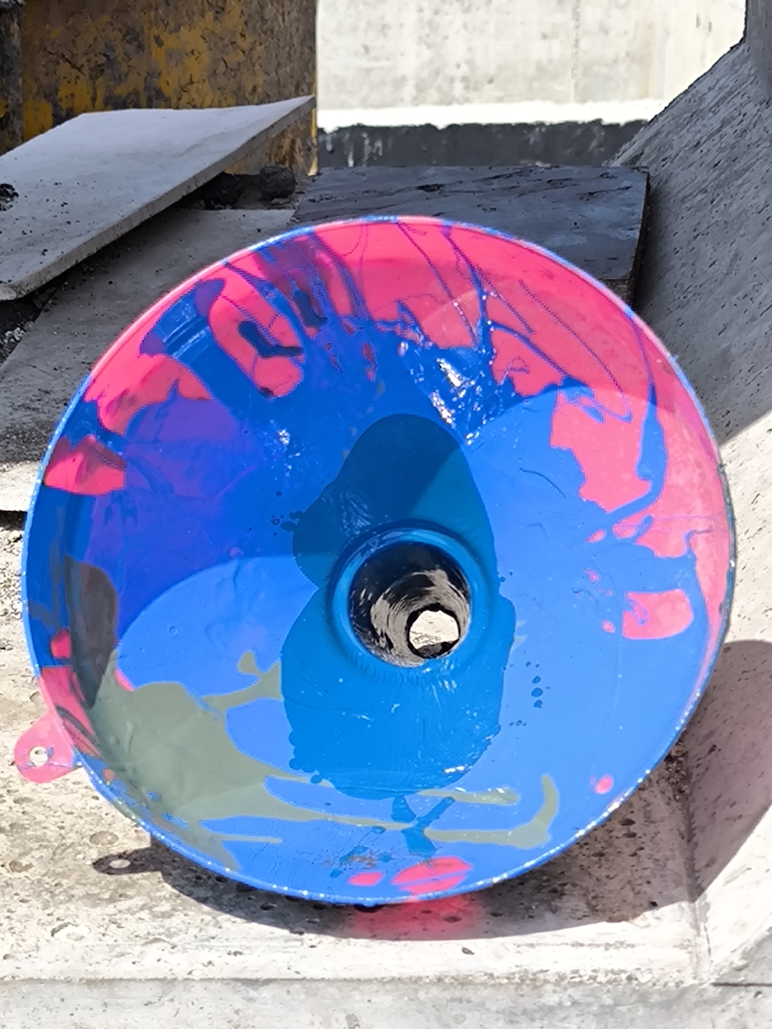 A low-angle, full-shot photograph shows a funnel with pink and blue paint inside and sitting on a textured concrete surface. The background includes other pieces of concrete debris and walls, suggesting a construction site or an area under renovation. The funnel's interior surface is primarily covered in blue paint, with areas of pink around the rim. The paint appears to be applied in a haphazard, splashed manner. The center of the funnel is open, revealing a dark interior. The concrete surface beneath the funnel is textured and gray, reflecting the ambient light. The background contains additional pieces of concrete, along with a concrete wall.
