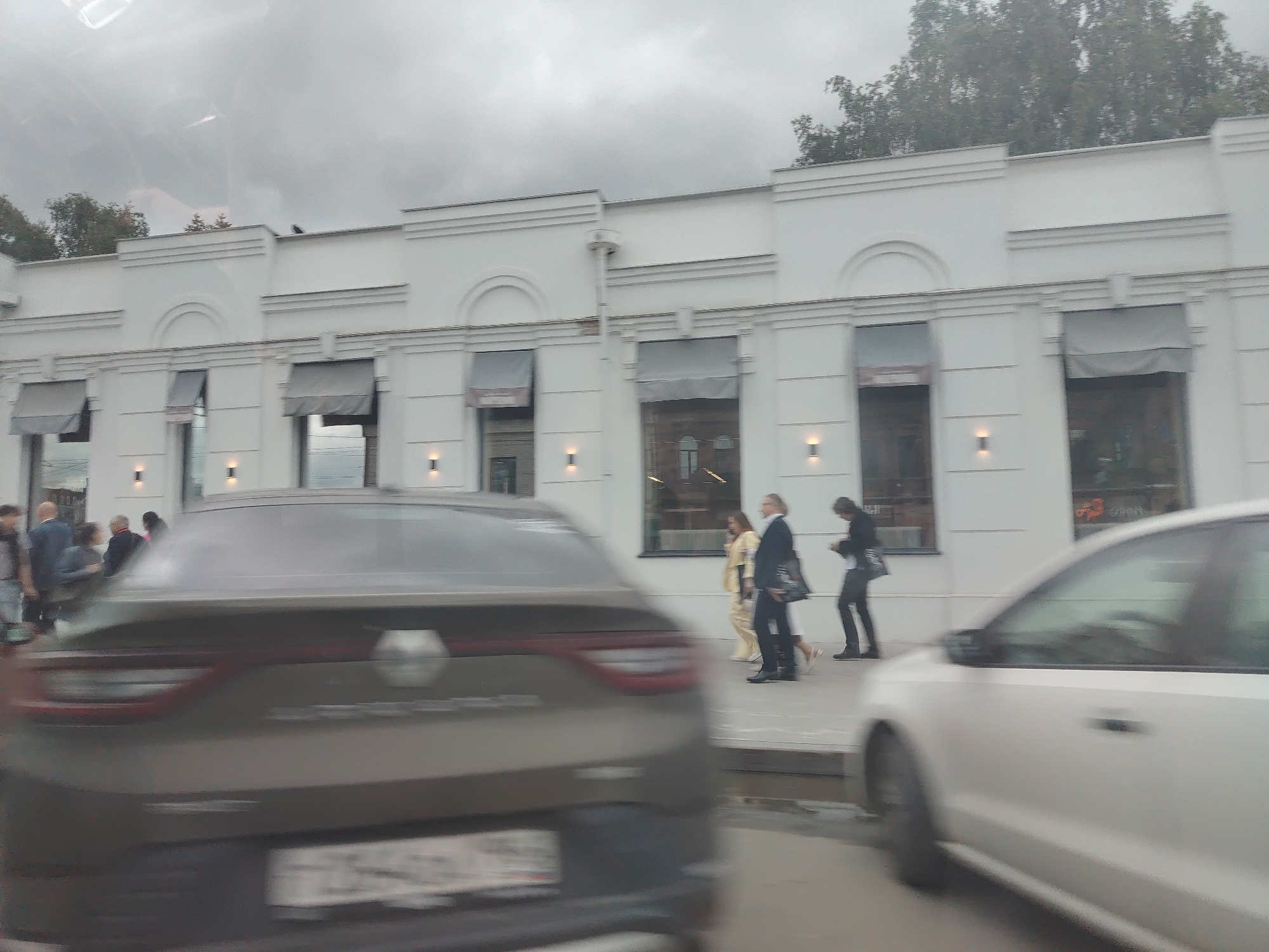 The image depicts a street scene in front of a modern white building with awnings over the windows. Several people are walking along the sidewalk. In the foreground, a dark-colored sedan is driving away from the camera, with its license plate blurred. To the right, a white car is partially visible. The weather appears overcast. The people visible include a couple walking together, a woman in a yellow outfit, and two men in suits. One of the men is carrying a bag. Another person is looking at their phone. The building has several windows with dark frames and what appear to be lights mounted below them. Trees are visible in the background, behind the building. The overall impression is a casual street scene with people on the move.