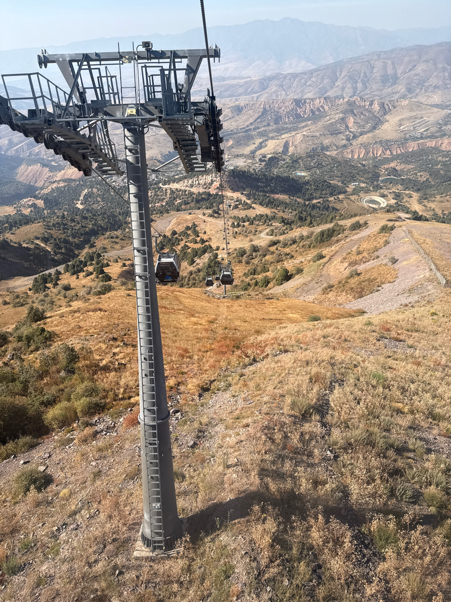 An aerial perspective captures a ski lift structure and several cable cars ascending a rugged, mountainous terrain in Nurekata, Uzbekistan. The ski lift's massive gray metal support tower dominates the foreground, with a ladder affixed to its side. Several dark-colored cable cars are visible, suspended from a thick cable that stretches across the frame. One cable car, marked with the word "AMIRSOY," is closer to the viewer. The landscape is characterized by dry, golden grasses, scattered green shrubs, and rocky inclines. In the background, distant, hazy mountains rise, their slopes carved by erosion into distinct formations. The scene suggests a sunny day with clear skies, the light casting long shadows across the terrain.