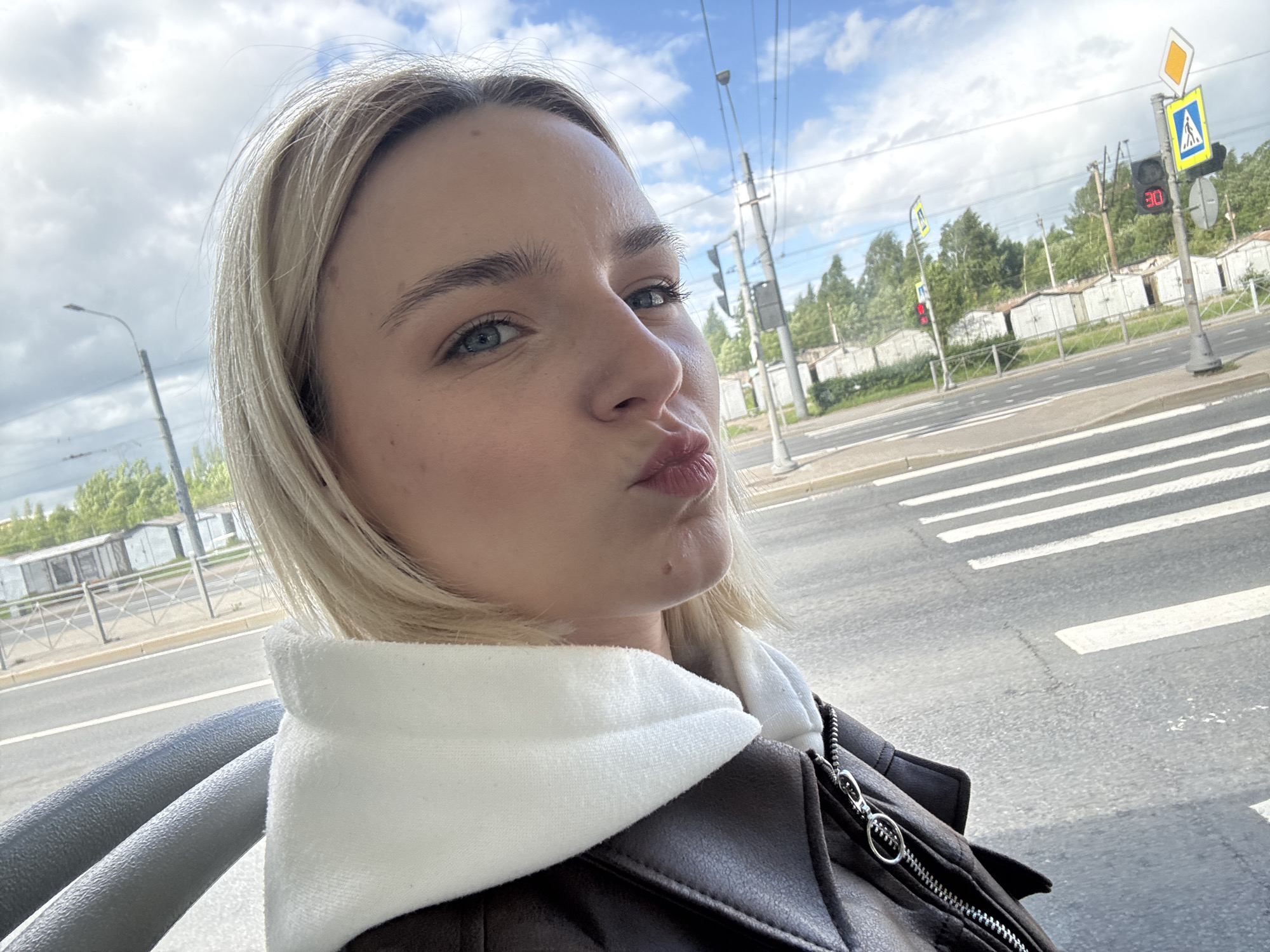 A young, fair-skinned woman with blonde hair and blue eyes is taking a selfie. She is puckering her lips in a kissing gesture. She wears a dark brown leather jacket with a white hoodie underneath. She appears to be sitting in a vehicle, possibly a bus or tram, with a gray handrail visible on the left. Through the window, a street scene unfolds in Saint Petersburg, Russia. There is a pedestrian crossing and a red traffic light displaying "30," presumably indicating 30 seconds. Behind the road, there are rows of garages and green trees. The sky is cloudy with patches of blue. A pedestrian crossing sign is visible. Overhead power lines are present.