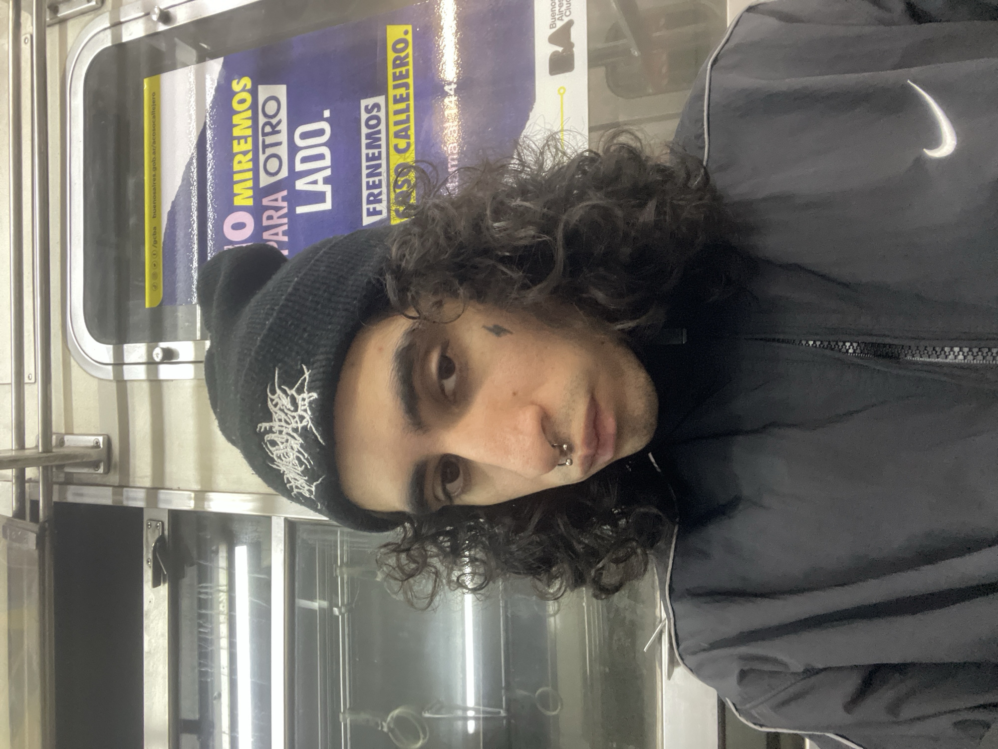 A medium shot depicts a person with dark curly hair, a beanie with indiscernible white text, and a black jacket with a white Nike logo. The person has a teardrop tattoo under their eye and a pierced septum. They are situated inside what appears to be a subway car or train, evidenced by the metal interior and a poster visible behind them. The poster features the phrases "O MIREMOS PARA OTRO LADO" and "FRENEMOS ACOSO CALLEJERO," along with text referencing "buenosaires.gob.ar/acosocallejero" and "Mamá al 144." There is also the Buenos Aires City logo. This indicates the location is likely Buenos Aires, Argentina, and the poster pertains to a campaign against street harassment.