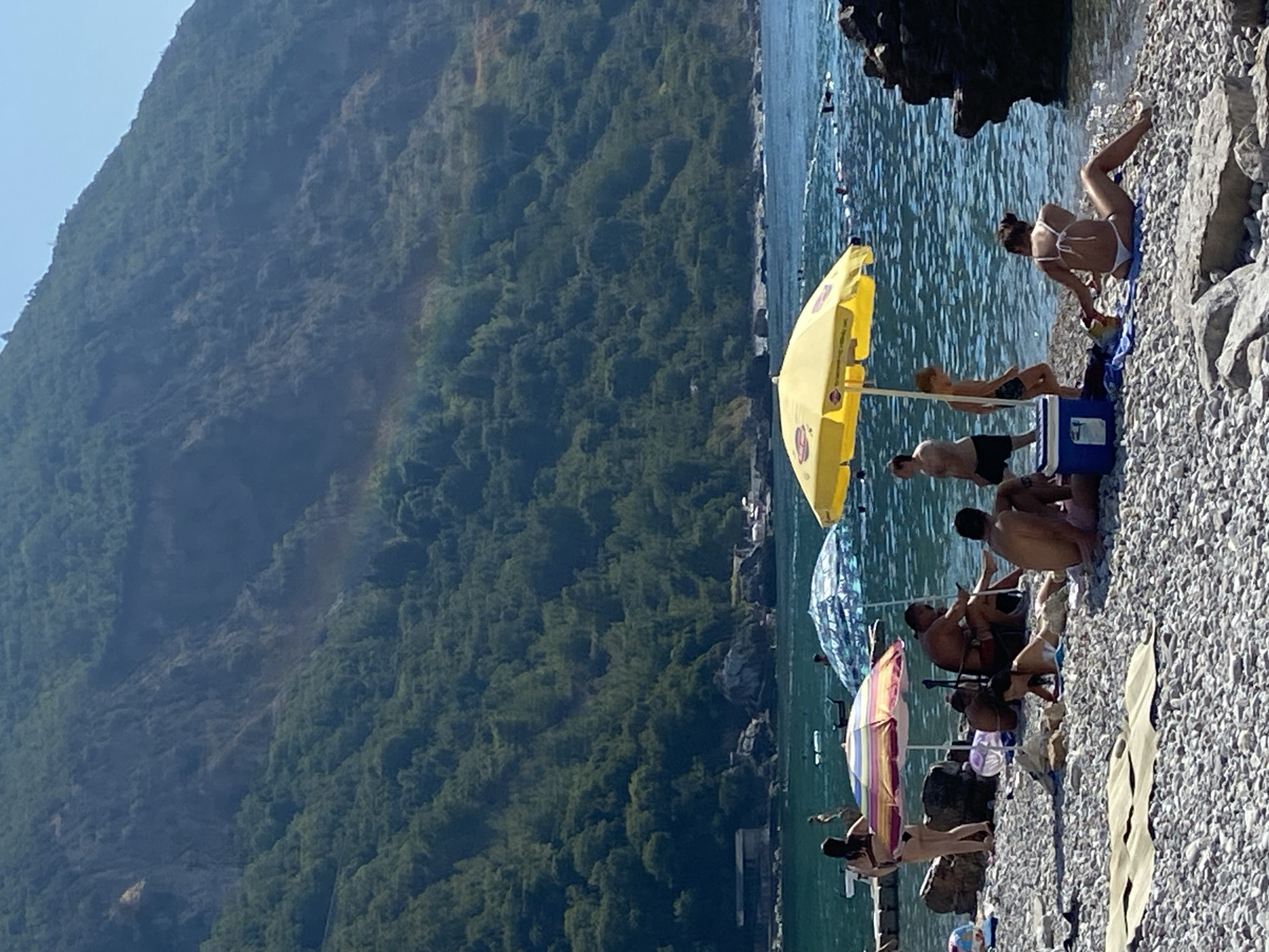 The image depicts a sunny day at a pebble beach in Prijevor, Montenegro, with a steep, tree-covered mountain rising behind the coastline. A small, faint rainbow is visible on the mountainside. The foreground shows several people relaxing on the beach. A yellow umbrella shades a portion of the beach, under which a man and a child are standing, with a blue cooler box nearby. To the left, a woman in a bikini is seated facing away from the camera, reaching for something on the ground. Another umbrella with colorful stripes is partially visible, with people sitting under it. In the water, several individuals are swimming or wading, and some are using flotation devices. A dark rock formation juts out into the water on the right side of the frame. The water is clear and blue, reflecting the bright sky. The overall scene suggests a leisurely day at the beach.
