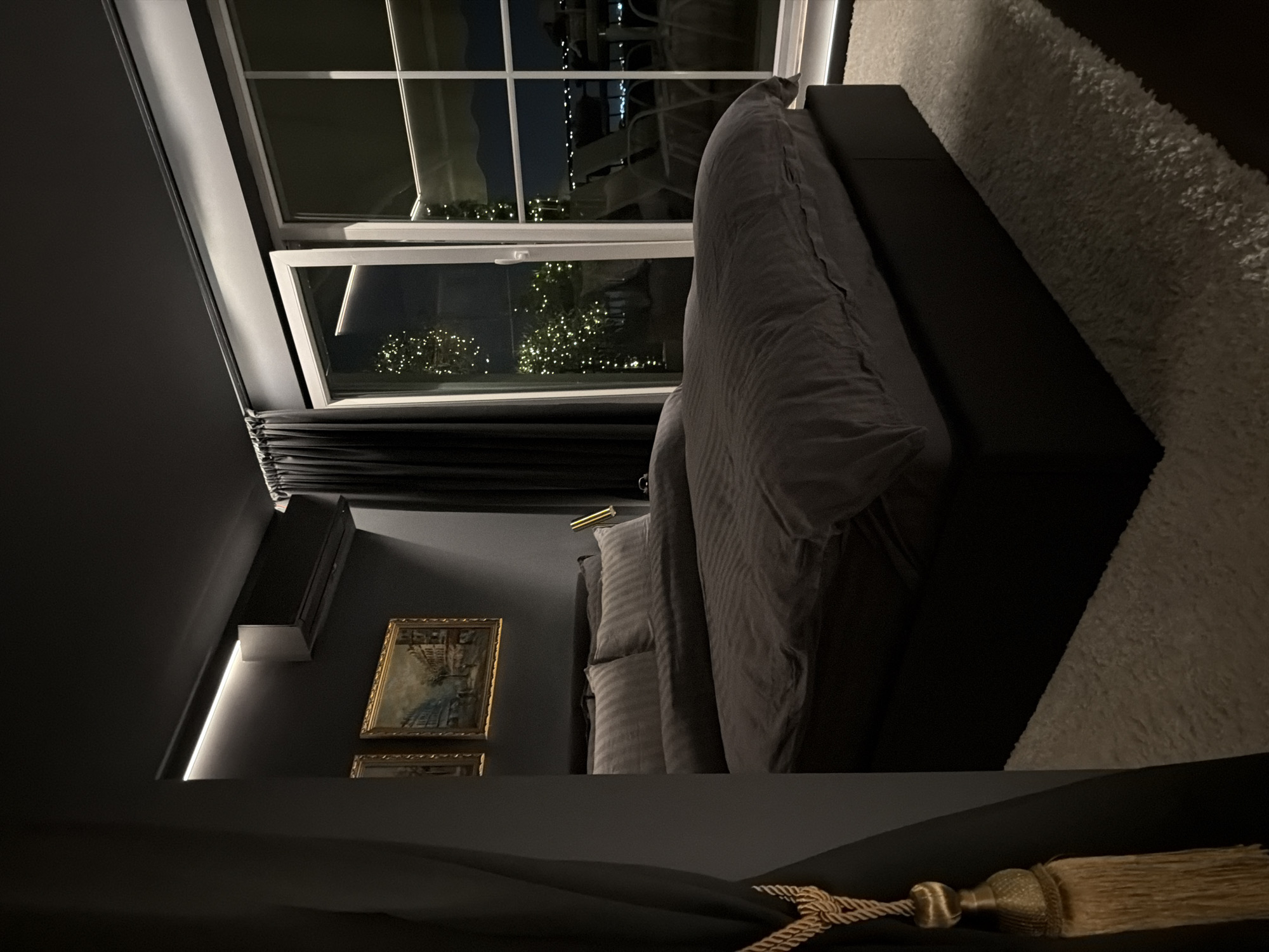 The image depicts a modern bedroom at night, likely in the city of Plovdiv, Bulgaria, given the view of the city lights and mountainous terrain visible through a large window. The room is dimly lit, with ambient lighting from the ceiling and string lights on an outdoor patio visible through the window. A large, dark-colored bed with gray striped bedding dominates the foreground. To the left, dark curtains with a gold tassel hang, partially obscuring a wall with two framed paintings. An air conditioning unit is mounted high on the wall above the paintings. The floor is covered by a plush white rug. The overall impression is one of a luxurious and contemporary urban dwelling.