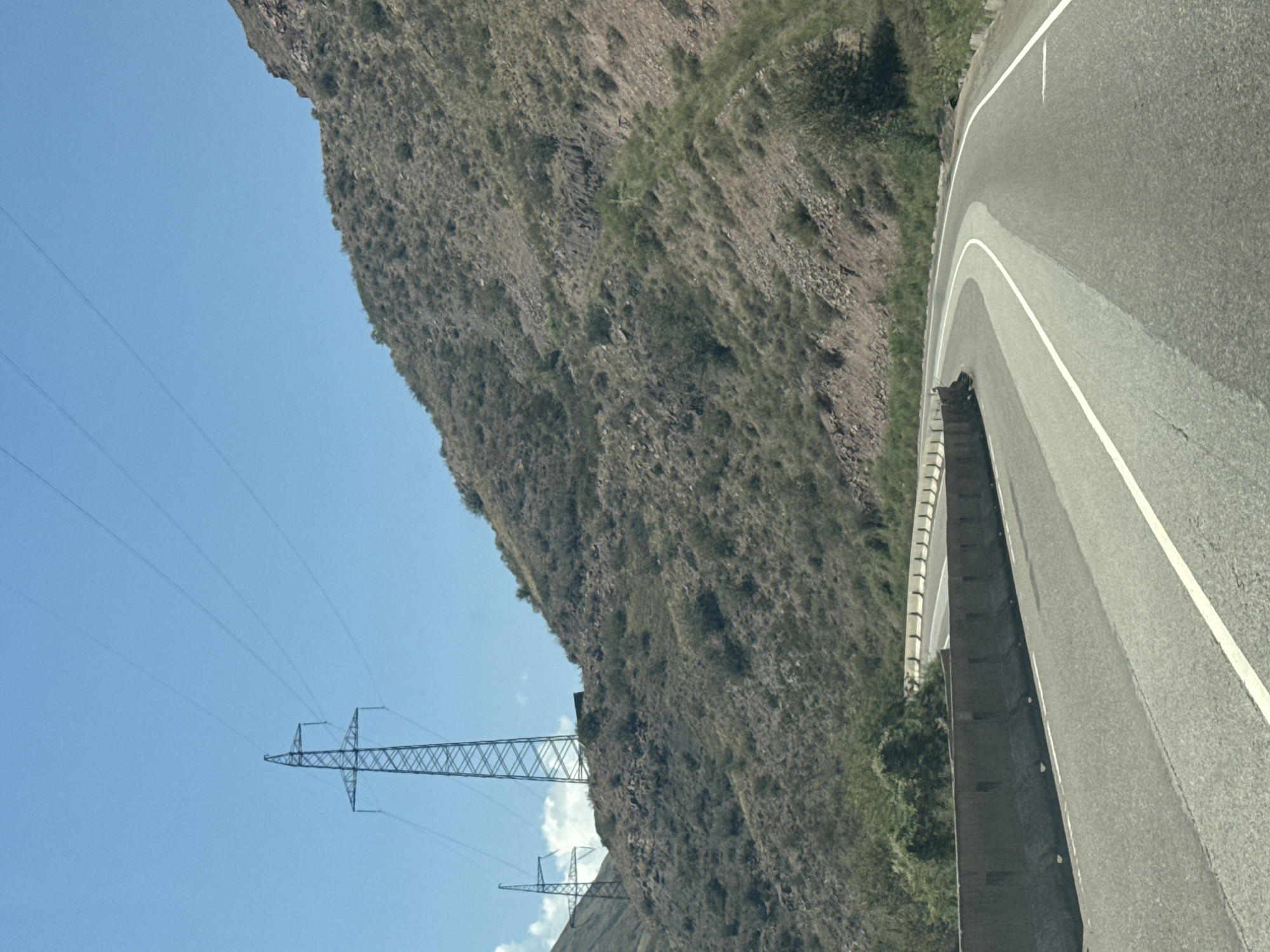 The image shows a road curving through a mountainous, arid landscape under a clear blue sky. Two tall power line pylons are visible on the left side of the frame, with power lines stretching across the sky. The road is paved and has white lane markings. A concrete barrier lines the left side of the road. The surrounding hills are covered in dry vegetation and rocks. The scene is devoid of people and any visible buildings or structures, except for the power line pylons. The overall impression is one of a remote, open highway in a natural environment. The image is likely taken from a moving vehicle, given the perspective of the road. The location is identified as the city of Cholok, Kyrgyzstan.