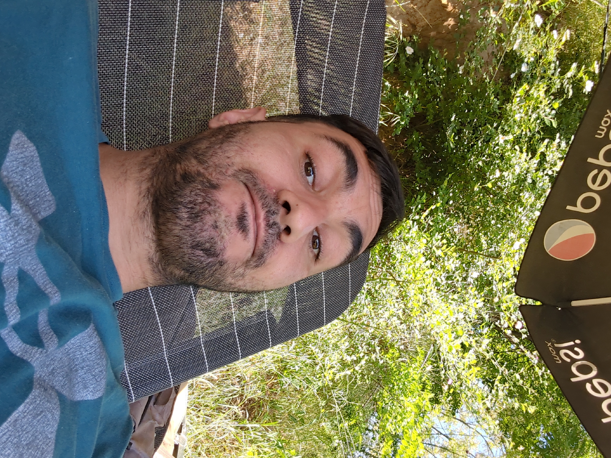 A medium shot of a fair-skinned man lying on a chaise lounge under a Pepsi umbrella. The man has dark hair and a beard. He is wearing a teal shirt with a grey graphic. The chaise lounge is dark grey and striped. Lush greenery surrounds the lounge. The location is presumed to be in Slavovitsa, Bulgaria.