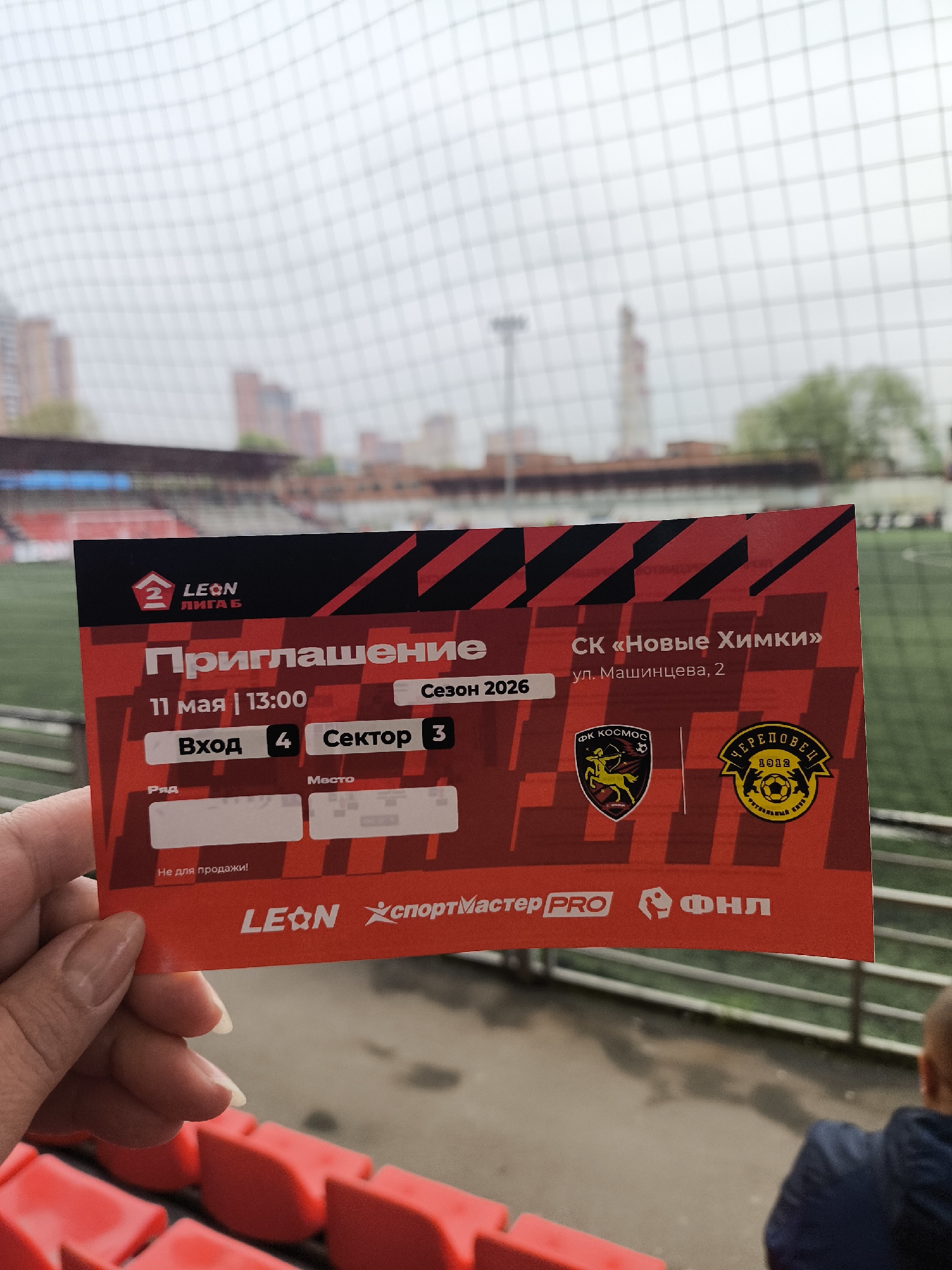 The image shows a person holding a ticket for a football match. The ticket, colored in red and black, is for a Leon Liga B game, season 2026, taking place on May 11th at 13:00. The ticket specifies entrance 4 and sector 3. The location of the event is "SK Novye Khimki", at 2 Mashintseva Street, in the city of Khimki, Russia. The ticket features the logos of "FK Kosmos", "Cherepovets 1912", "Leon", "Sportmaster Pro", and "FNL". In the background, a football stadium can be seen with red seating and a tall building in the distance. A mesh fence is also visible between the viewer and the stadium.
