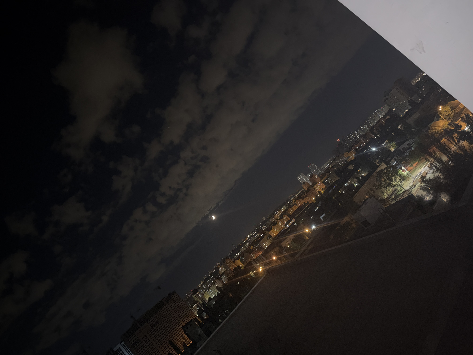 This is a night-time, high-angle, wide shot of a city skyline, angled slightly from left to right. The sky is dark with scattered clouds, some of which are illuminated by the city lights below. A crescent moon is visible in the upper right quadrant of the sky. The cityscape below is densely populated with buildings of various sizes and heights, illuminated by numerous artificial lights. Streetlights and lights from within buildings create a warm glow across the urban landscape. In the foreground, a dark, smooth surface, possibly a balcony railing or wall, takes up the bottom left portion of the image. The overall impression is of a bustling city at night under a cloudy sky, with a clear view of the moon. The location in Kyiv, Ukraine is not discernible from the image itself. There are no people or discernible actions depicted in the scene.