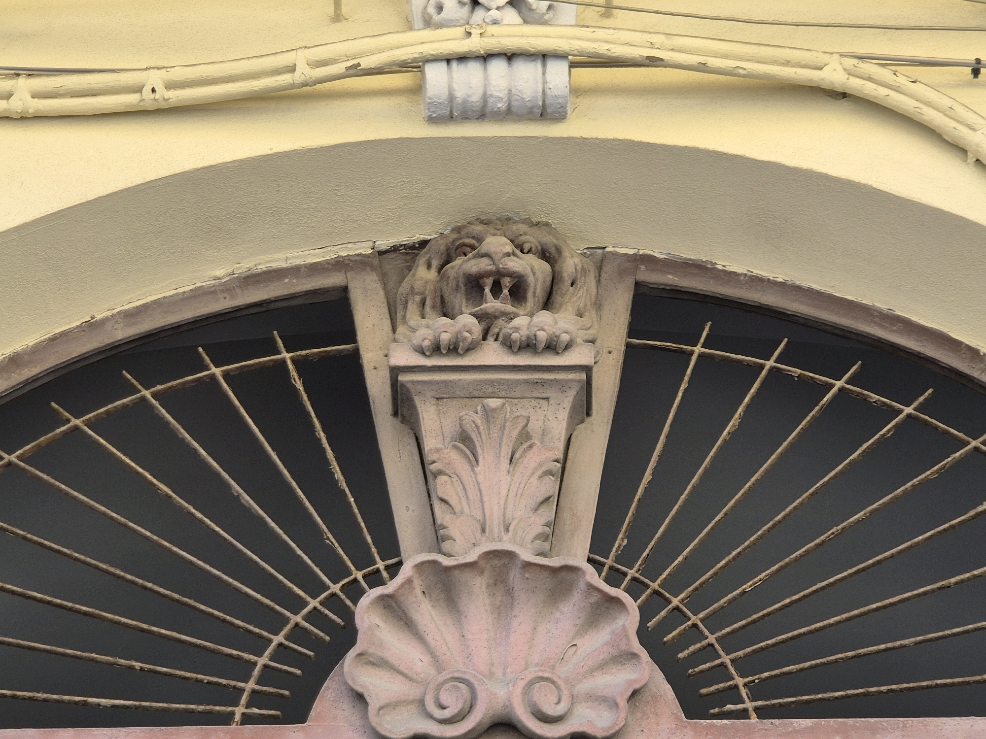 An arched window, partially obscured by a radial metal grate, is adorned with a stone carving of a lion's head. The lion's head carving protrudes from a decorative stone pedestal, which features acanthus leaf and shell motifs. Above the arch, a yellow-painted building facade exhibits some peeling paint and external conduits. The architectural style suggests a building in a historical urban setting. This image was captured in Taranto, Italy.