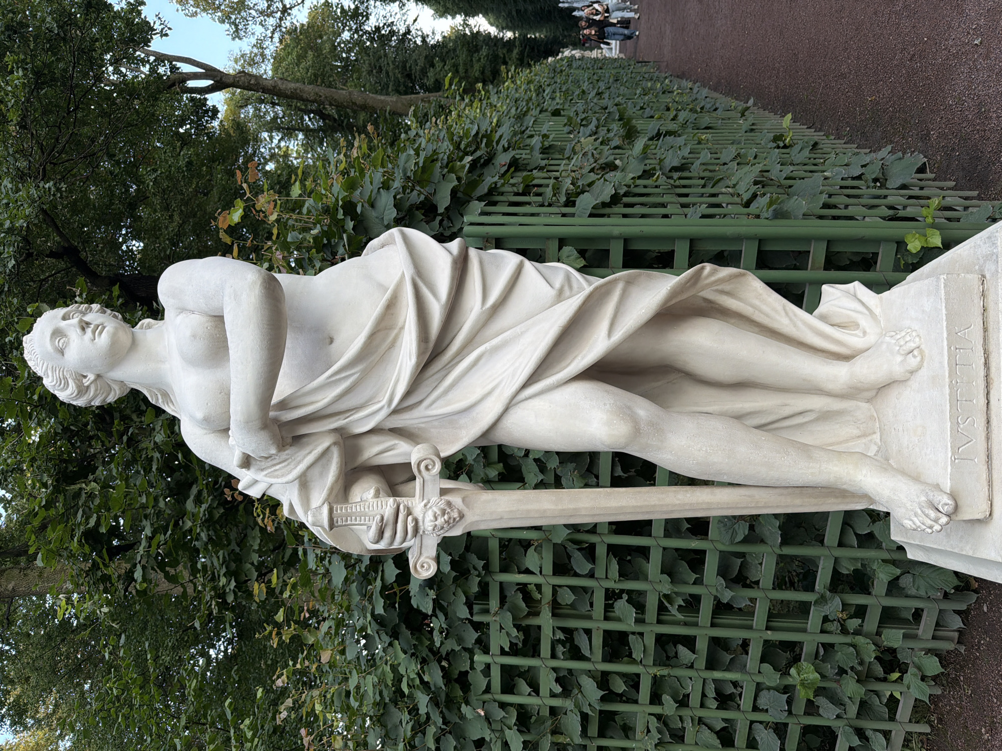 The image shows a statue of the Roman goddess Justitia in the Summer Garden, Saint Petersburg, Russia. Justitia, identified by the inscription "IVSTITIA" on her pedestal, is depicted as a semi-nude female figure draped in classical robes, holding a sword in her left hand. The statue is made of white marble and stands on a grey stone pedestal. It is positioned in front of a green lattice fence overgrown with ivy, with lush green trees in the background. To the right of the statue, a path lined with more green lattice fencing and trees leads into the distance, where several people are walking. The path is covered with dark reddish-brown gravel. The overall atmosphere is serene and classical, typical of a European garden setting.
