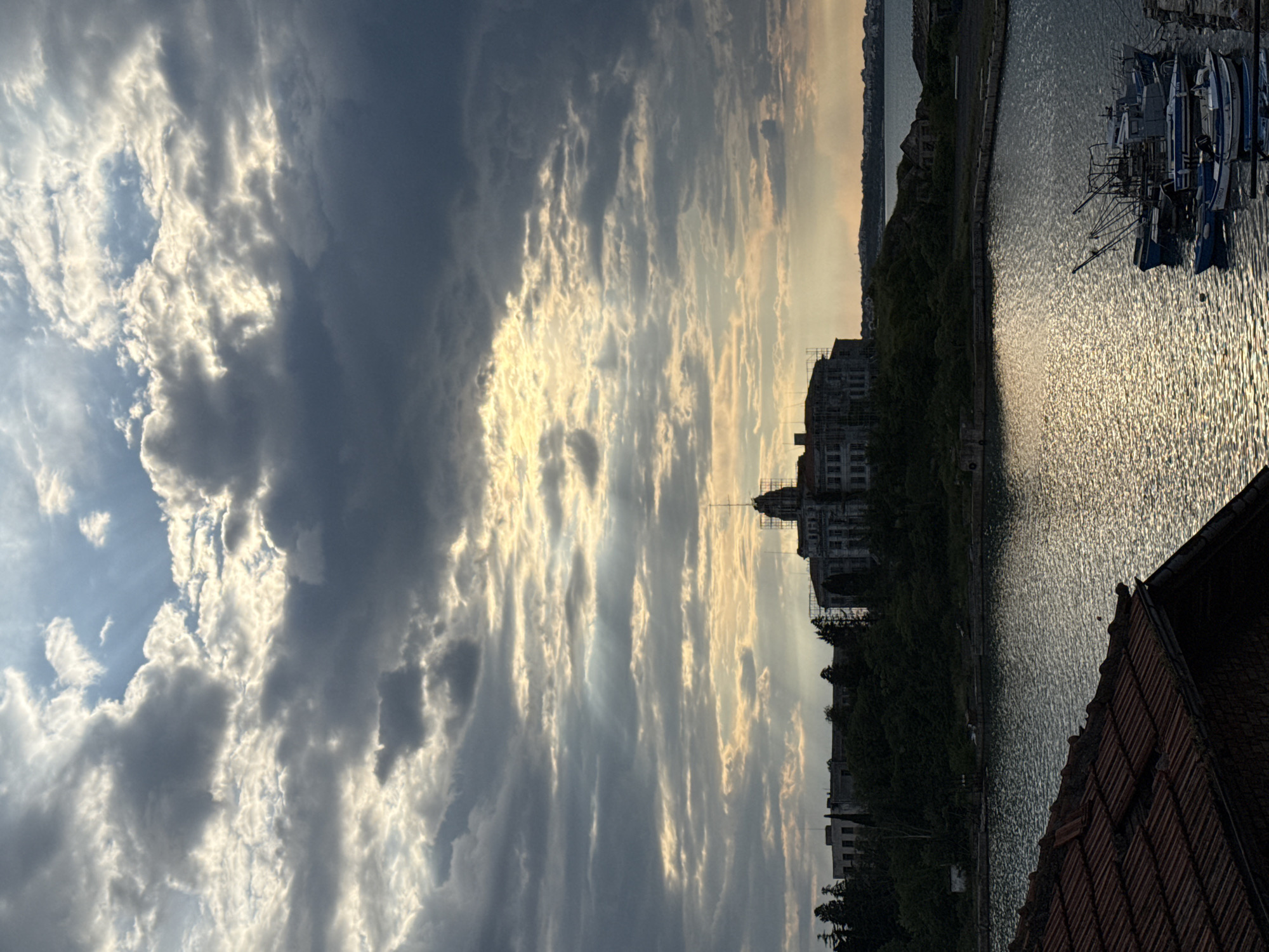 The image shows a cloudy sky over the Bulgarian coastal city of Sozopol. A building is under construction, with scaffolding visible around it. The water appears to be shimmering, reflecting light. Several boats are docked in the water. In the foreground, there is part of a tiled roof.