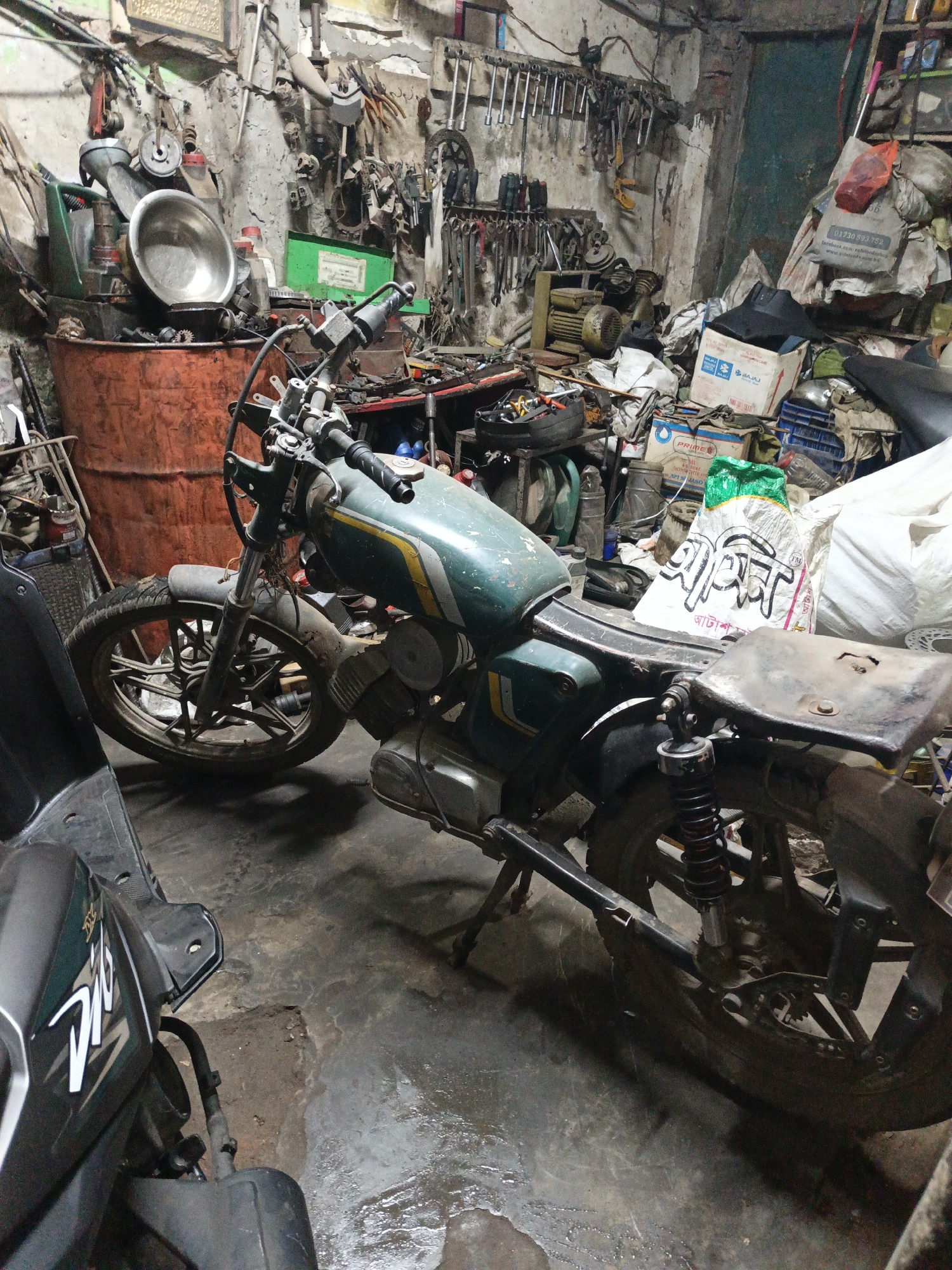 The image depicts a cluttered motorcycle repair shop, likely in Dhaka, Bangladesh. Two motorcycles dominate the foreground: one an older, teal and yellow model with visible wear and tear, resting on its kickstand, and the other, a partially visible, modern black scooter marked "DIO". The floor is wet and stained, suggesting frequent use and spills.
In the background, the shop is filled with tools hanging on the walls, parts scattered on tables, a large orange barrel, and boxes of goods. A partially open door is visible in the upper right. The overall environment is cramped and busy, indicative of a small, independent repair operation. The tools and spare parts suggest the shop is actively engaged in motorcycle maintenance and repair. The presence of the bag with text in Bengali script further corroborates the location as Bangladesh. There are no people visible in the frame.