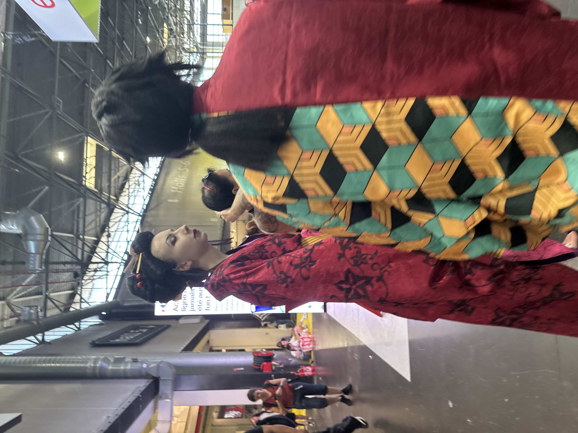 Two individuals are in the foreground of a well-lit indoor event space, likely a convention center. The person on the left, viewed from the side, is wearing a vibrant red kimono with black floral patterns and has an elaborate updo hairstyle with red accents. This person has dramatic eye makeup. The person on the right is viewed from behind and wears a garment that is half plain red and half patterned with a geometric design in teal, yellow, and black. They have dark hair styled in a ponytail that extends down their back. In the background, other attendees are visible, some browsing displays or walking. Signage for "Halls 5-6" is present on a wall in the mid-ground, indicating this is likely the Paris-Nord Villepinte Exhibition Centre in Villepinte, France, a common venue for large-scale events and conventions. The floor is a light gray, and the ceiling features exposed industrial beams and ventilation systems.