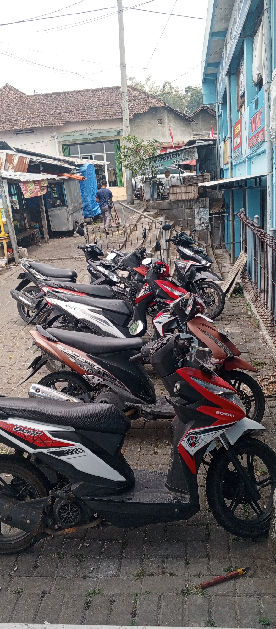 The image shows a row of parked scooters in front of a building that appears to be a shop or market stall in Plaosan, Indonesia. The building has a blue facade and a metal fence. There are several scooters parked in a line, most of them are red and black. One scooter in the foreground is white and black with red accents and the word "Beat" on its side. A person wearing a blue shirt and grey pants is walking away from the viewer towards the building. The ground is paved with cobblestones. In the background, there are other buildings with tiled roofs, and some vegetation. There is also a small cart with a blue tarp visible. The overall scene depicts a typical street view in a town in Indonesia.