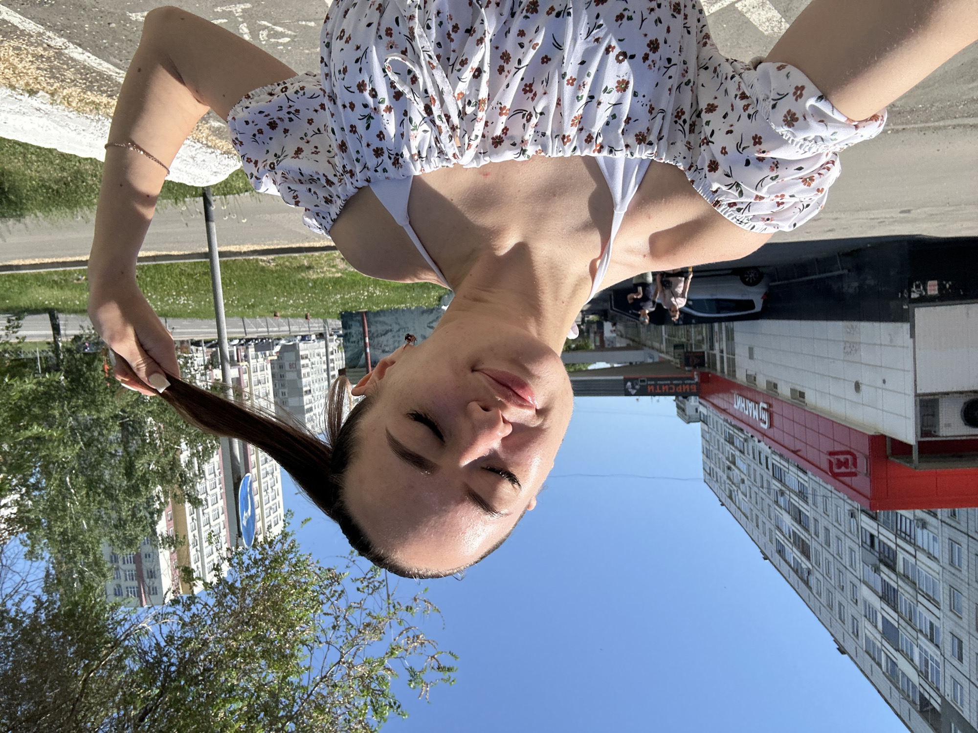 The image depicts a young adult female in the foreground, taking what appears to be a selfie on a clear, sunny day in an urban environment. She is shown from the chest up, with her head tilted slightly, eyes mostly closed, and a subtle smile. Her dark hair is pulled back into a ponytail, which she holds with her right hand. She wears a white off-the-shoulder top adorned with small brown, orange, and green floral patterns, layered over a white halter-neck garment. A delicate, light-colored bracelet is visible on her left forearm.
The background reveals a city street scene. To the left, a tall, multi-story grey residential building dominates the skyline. Below it, a red commercial facade prominently displays the Cyrillic word "МАГНИТ" (Magnit) and a distinct "M" logo in white. A white commercial structure and an air conditioning unit are visible beneath the red facade. A silver hatchback car is parked on the street, and two indistinct figures are walking further back on the left side. To the right of the subject, green trees with dense foliage are interspersed with more multi-story residential buildings in the distance, some featuring sections of yellow and pink facades. A blue circular road sign with a white right-pointing arrow is visible among the trees. The foreground consists of a paved surface, likely a road or sidewalk, with a white curb or low wall to the right. The sky above is a clear, bright blue. Cyrillic script is present on the commercial signage.