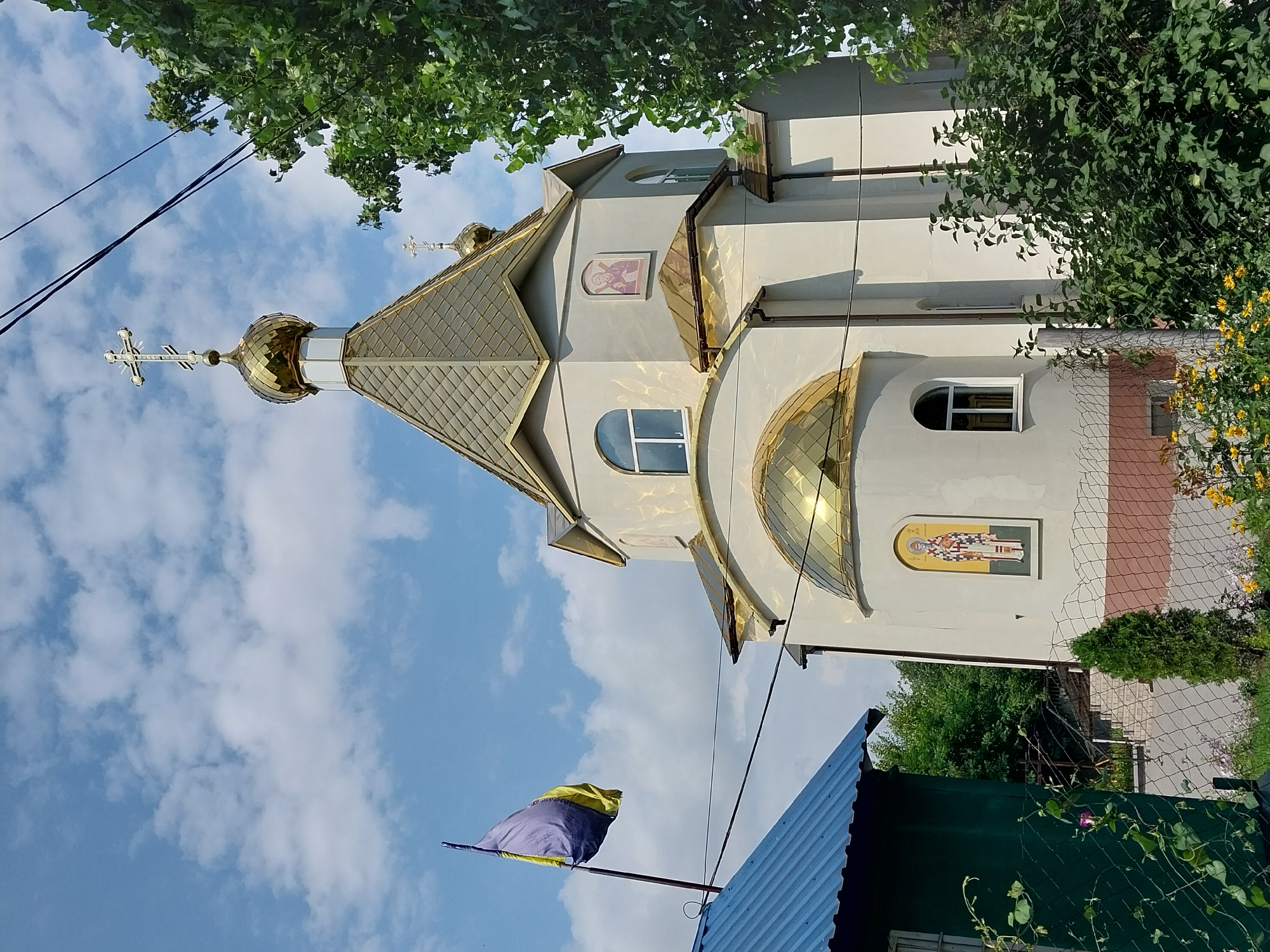 The image shows an Orthodox church with a golden dome, located in the city of Doslidne, Ukraine. The church is white with arched windows and decorative religious icons on its exterior. A Ukrainian flag is visible on a flagpole to the left of the church. In the foreground, there is a green building with a blue roof, a chain-link fence, and vegetation including flowers and trees. The sky is blue with scattered white clouds. The scene is captured from a slightly low angle, looking up towards the church and the sky.