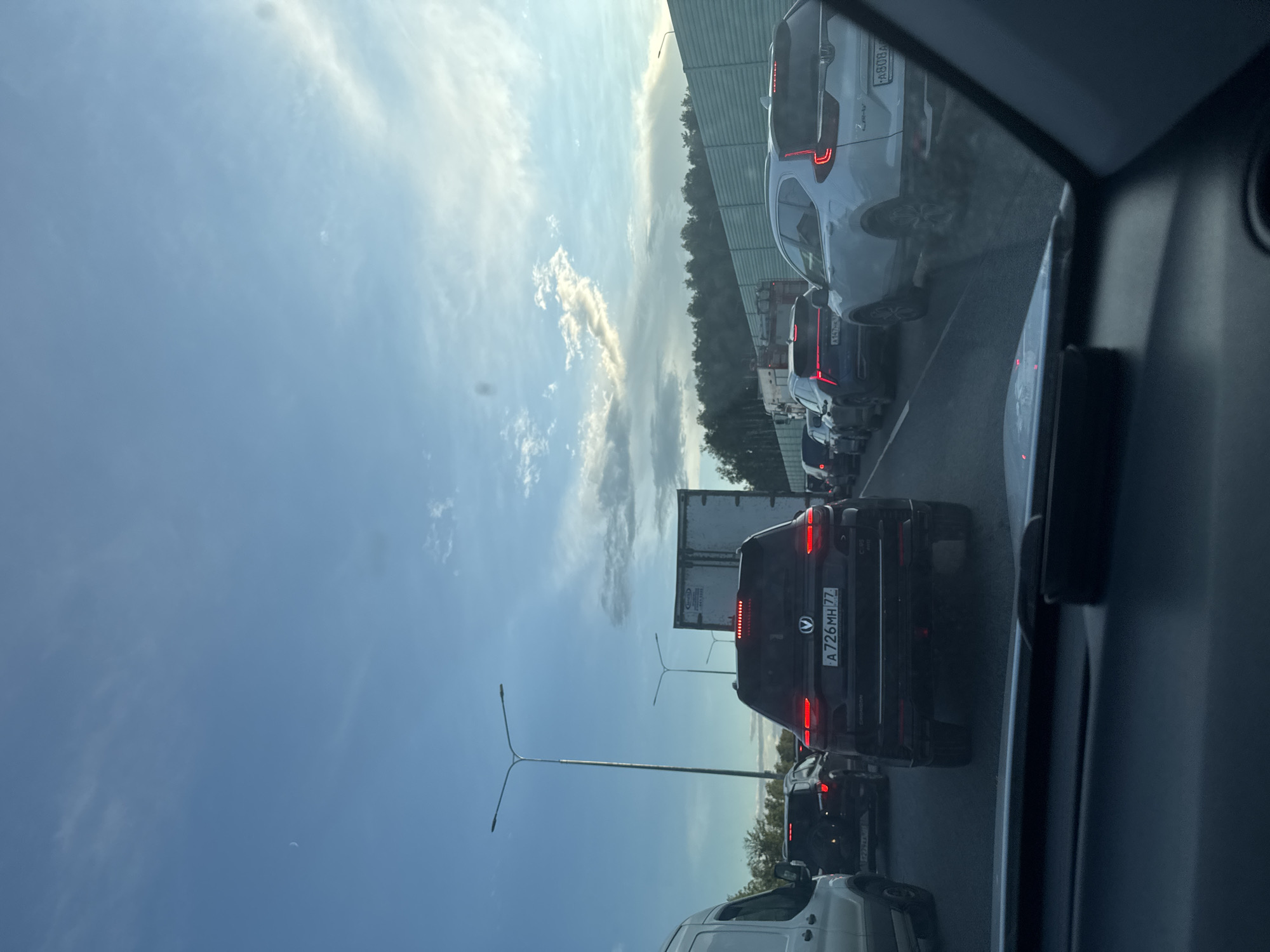 The image depicts a traffic jam on a highway, likely near Krutitsy, Russia, given the presence of Russian license plates. The perspective is from inside a vehicle, looking forward. Several cars are visible in multiple lanes, with brake lights illuminated, indicating slow or stopped movement. The sky is a clear blue with scattered clouds and a crescent moon. Trees and a sound barrier line the side of the highway. Streetlights are also present. The overall scene suggests a typical urban or suburban commute during daylight hours.