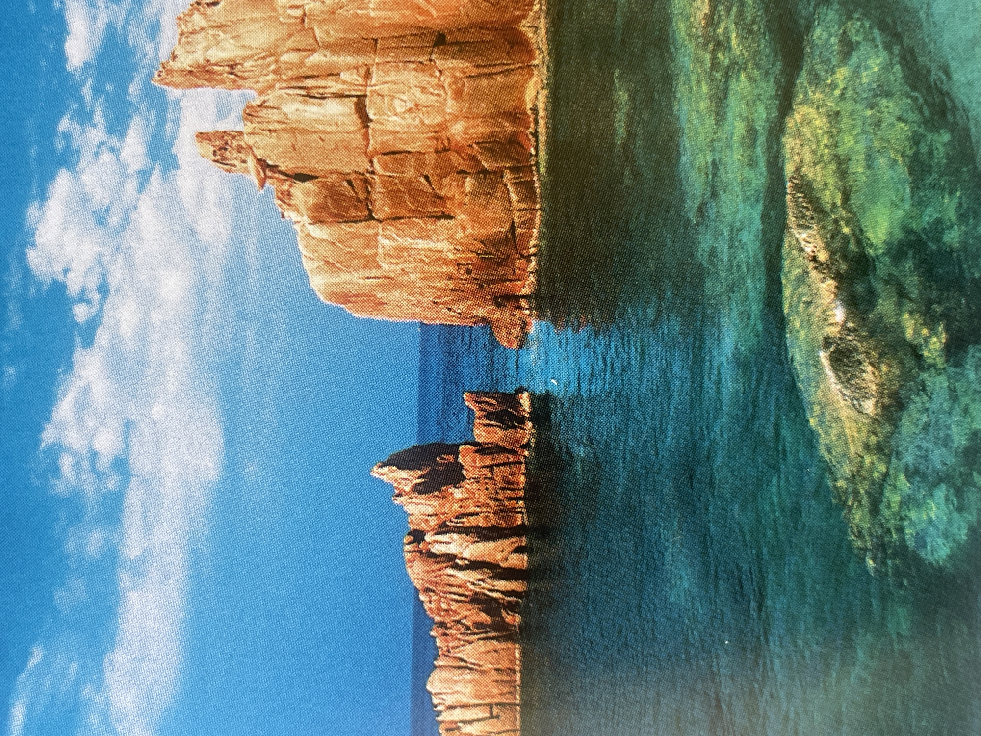 The image displays a scenic view of jagged, reddish-brown rock formations emerging from clear, turquoise water. The sky above is a vibrant blue with scattered white clouds. The rock formations appear weathered and have distinct vertical and horizontal lines, suggesting sedimentary layers. One larger formation is on the right side of the image, and a smaller, more fragmented formation is on the left. In the foreground, underwater rocks or coral are visible beneath the surface of the water, illuminated by sunlight. The overall impression is a natural, coastal landscape. This image does not depict the city of Sofia, Bulgaria.