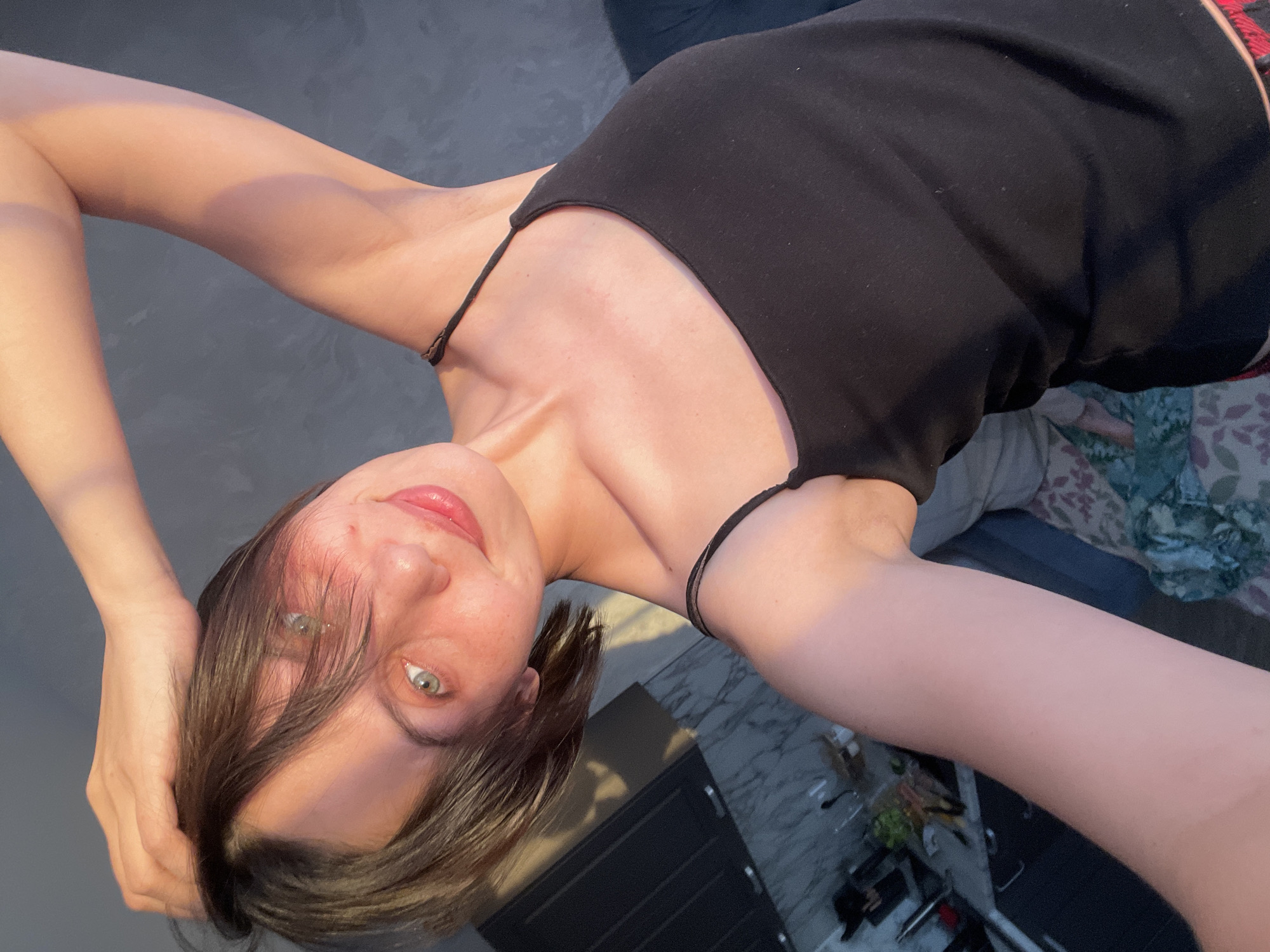 A selfie of a woman with dark brown hair and fair skin, wearing a black tank top. Her right arm is raised, with her hand touching her head. The background includes a gray floor, a partial view of a bed with patterned bedding, and a glimpse of other furniture. The location within Izhevsk, Russia cannot be determined from the provided image.