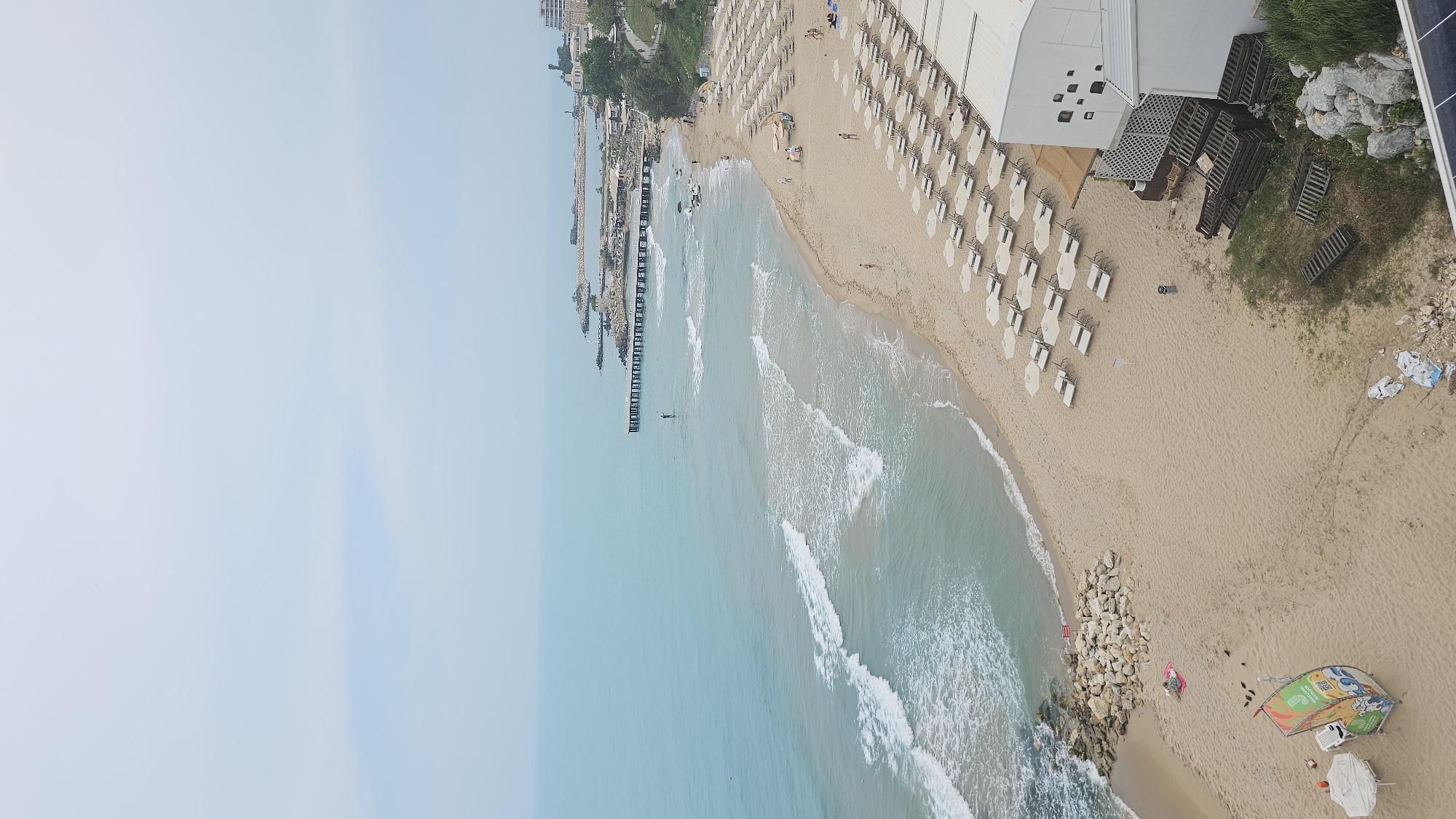 This aerial photograph presents a serene coastal scene in Varna, Bulgaria, offering a high-angle view of a sandy beach, the sea, and adjacent infrastructure. The image shows a wide stretch of golden sand, meeting the clear, light blue waters of the sea. Gentle, white-capped waves rhythmically break along the shore.
On the beach, numerous rows of light-colored beach umbrellas and sun loungers are neatly arranged, predominantly towards the upper right and mid-right sections, indicating an organized resort or public beach area. Most of these appear unoccupied, suggesting a relatively uncrowded day. Several individuals are scattered across the sand, some relaxing or sunbathing, while others are seen near or in the shallow waters.
Towards the middle-left of the image, a long, dark pier extends into the sea, flanked by rocky breakwaters. Beyond the pier, the coastline curves, revealing more distant buildings and green areas. A few small, dark objects, possibly buoys or small boats, are visible in the water.
On the land side, specifically in the upper right, a modern white building, likely a beach club or restaurant, stands behind a section of beach furniture. Adjacent to this building is a patch of green grass with large rocks and stacks of wooden pallets. In the bottom right foreground, a distinctive, colorful structure resembling a partially buried boat or a large, painted signboard with cartoonish graphics and some indistinct text (including visible letters like 'E' and 'N') adds a focal point. A person is seen lying on a pink towel near this structure. A portion of a solar panel is also visible in the top right corner.
The sky is a pale, uniform blue, indicating clear weather. The lighting suggests it is daytime, possibly late morning or early afternoon, with a generally calm and relaxing atmosphere.