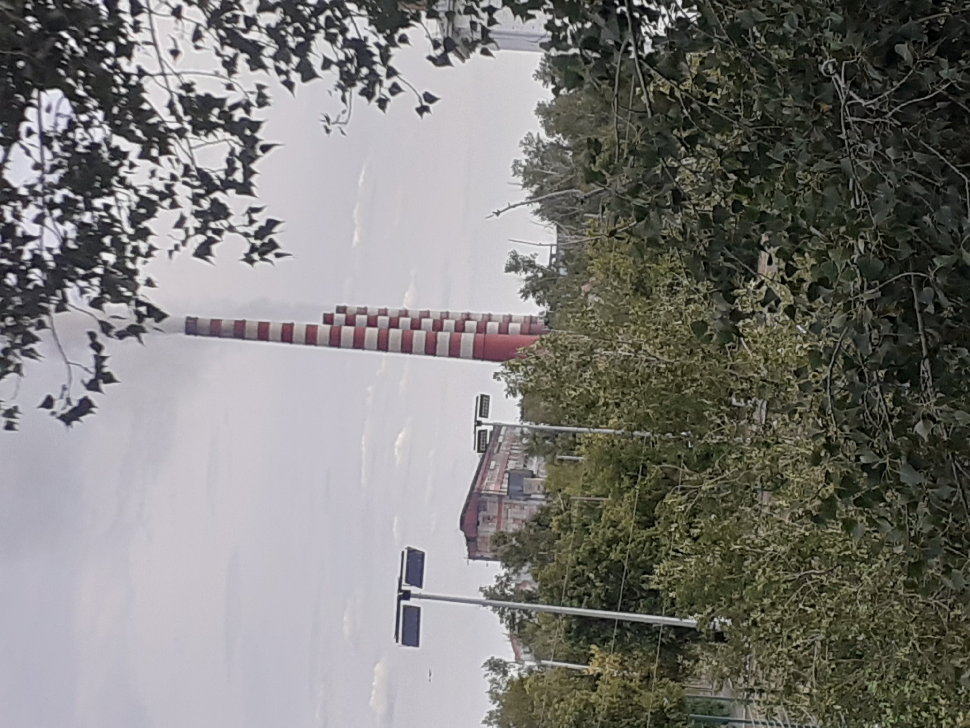 The image depicts an industrial area with a prominent, tall, red and white striped smokestack emitting smoke into a cloudy sky. In the foreground, a dense collection of green trees and foliage partially obscures the view. Behind the trees, a large, weathered industrial building with a red roof is visible, and two tall, modern streetlights with rectangular lamps stand out. The overall scene suggests an industrial zone within or near the city of Aksu, Kazakhstan, characterized by industrial infrastructure and surrounding vegetation.