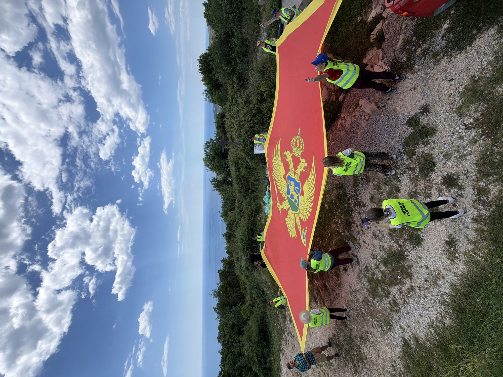 A group of people are holding a large Montenegrin flag on a hillside. The flag is predominantly red with a yellow border, and it features the coat of arms of Montenegro at its center.
People, wearing yellow vests, are positioned along the edges of the flag, holding it up. Some individuals are looking towards the camera, while others appear to be adjusting the flag. The terrain is a mixture of dirt, rocks, and green vegetation. There is a red car is partially visible.
In the background, there is a panoramic view of the sea and sky. The sky has numerous white clouds against a blue backdrop. Green trees and shrubs cover a portion of the hill.
The location is Budva, Montenegro.