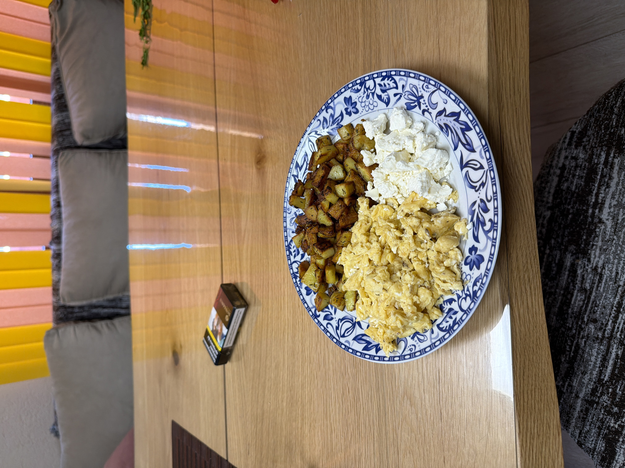 The image depicts a plate of food on a wooden surface, likely a table. The plate contains three distinct food items: fried potatoes, scrambled eggs, and feta cheese. A pack of cigarettes is placed on the table next to the plate. In the background, there is a couch with cushions and yellow blinds. The photograph could have been taken in Panagyurishte, Bulgaria, but there is no information to confirm that.