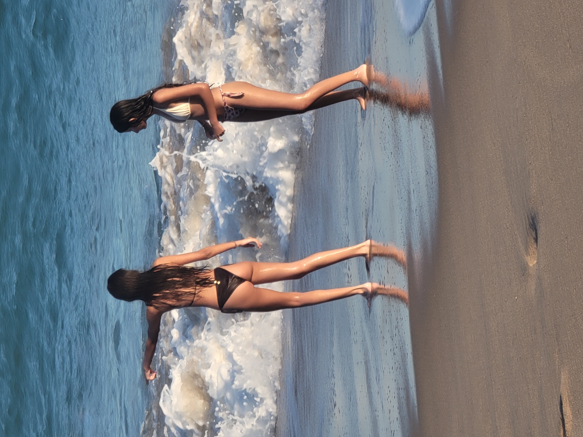 Two young women are lying on their stomachs at the edge of the ocean in Quarteira, Portugal. The woman in the foreground is wearing a black bikini, and the woman in the background is wearing a white bikini with a patterned bottom. They are both facing away from the camera, with their legs extended towards the sand. Gentle waves are washing over their legs, creating a frothy spray. The sun appears to be low in the sky, casting long shadows on the wet sand, suggesting it is either early morning or late afternoon. The ocean is a deep blue, and the sky is clear. The scene evokes a sense of relaxation and enjoyment of a day at the beach. There is no visible text in the image.