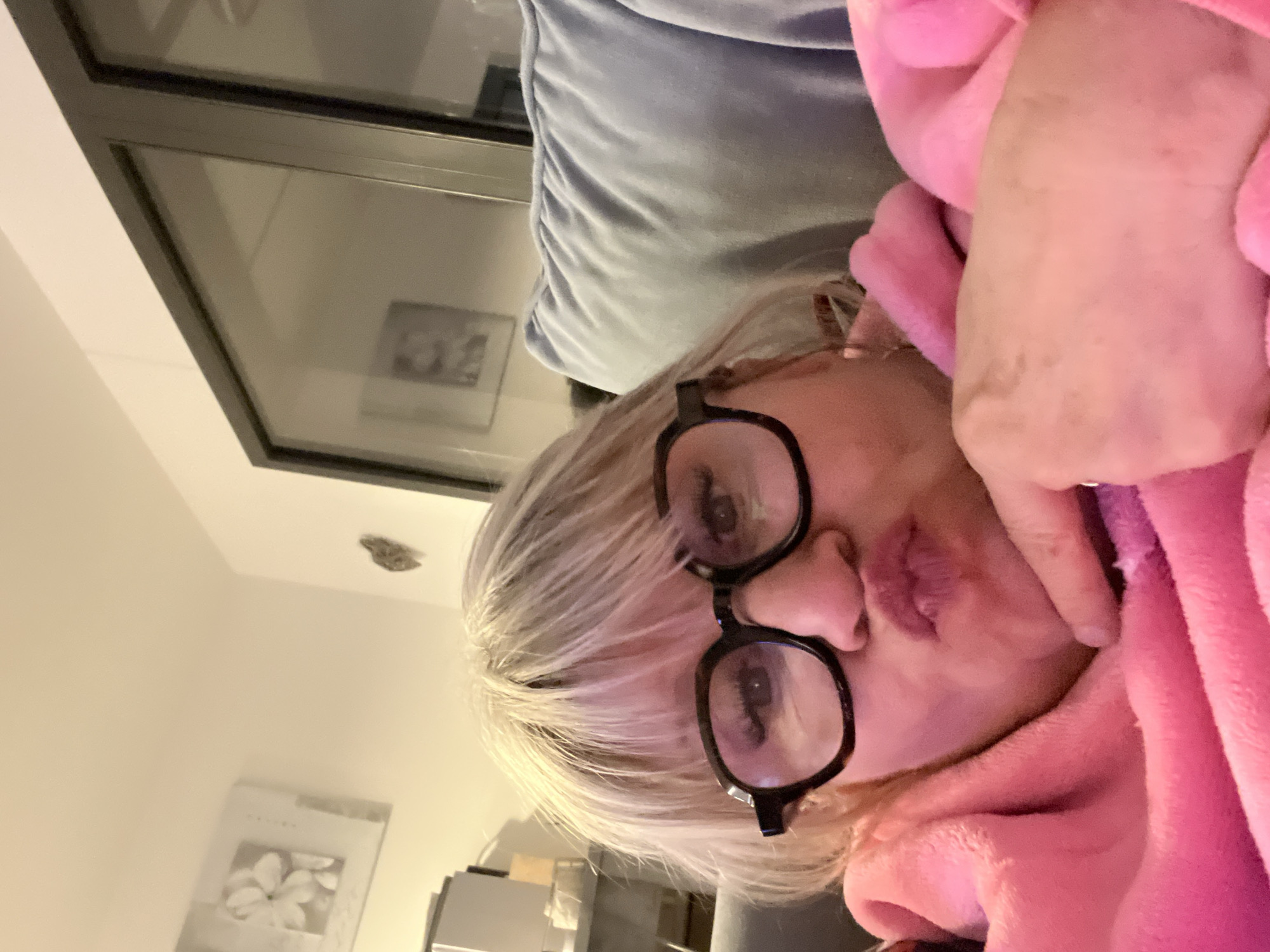 A selfie depicts a woman with light blonde hair, wearing black-rimmed glasses and a pink fuzzy robe, posing for the camera. The background includes a light blue pillow and a window with a dark frame. Reflections in the window suggest an interior setting with framed art on the wall. A white wall is visible, with a framed floral picture. The presence of a light fixture on the ceiling, possibly a vent, can be seen. There is no information to support the location is Chambœuf, France.