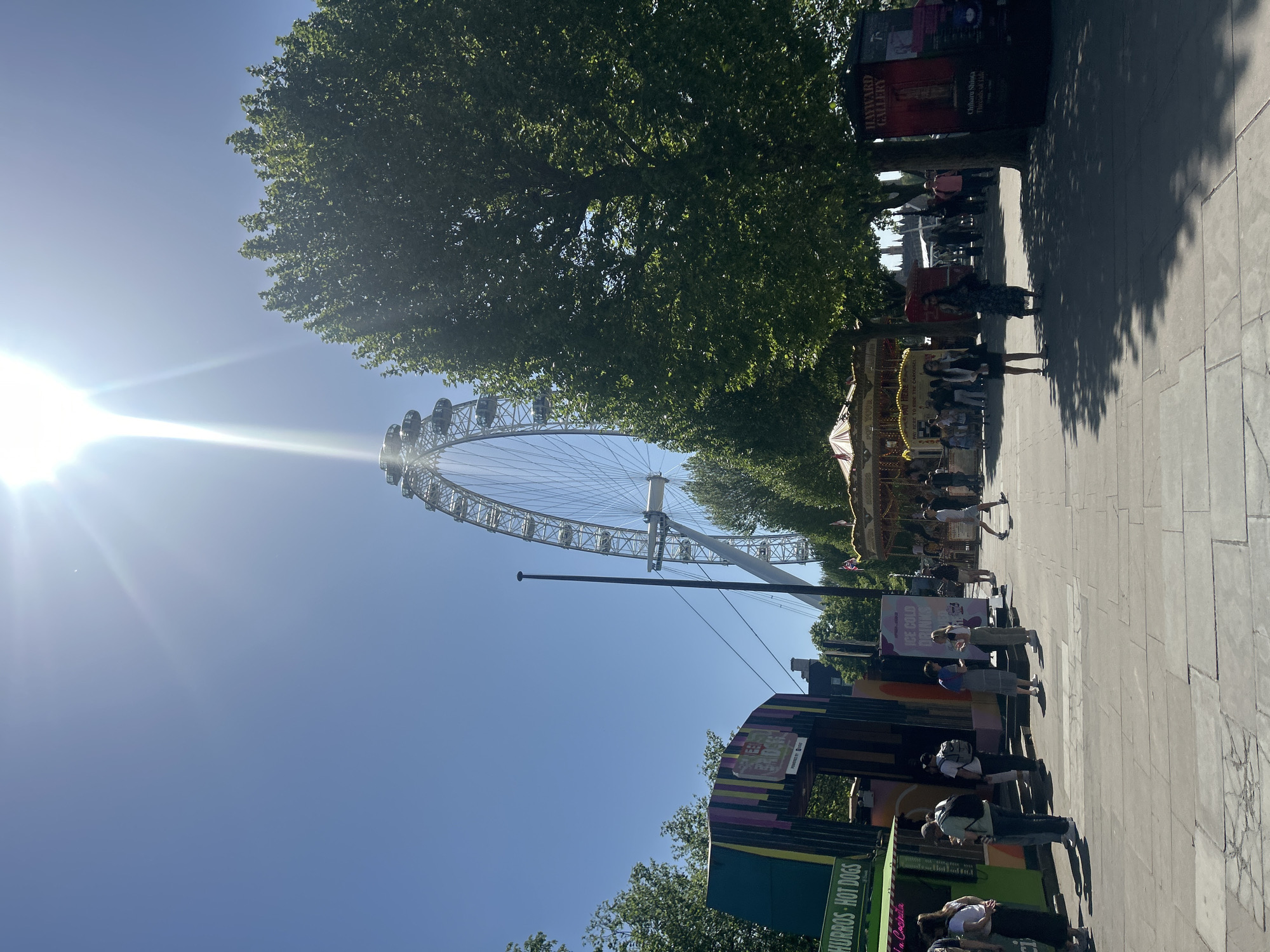 This image captures a vibrant outdoor scene on a bright, sunny day, likely along the South Bank in London, United Kingdom, given the prominent London Eye.
The main subject is the iconic London Eye, which dominates the upper left portion of the frame, silhouetted against a clear, brilliant blue sky. A strong sun flare emanates from the upper left corner, indicating direct sunlight and casting a bright glow. Lush green trees line the right side of a paved pedestrian pathway, their foliage providing shade.
The scene depicts a bustling public space, with numerous people walking and standing along the pathway. Their distinct, elongated shadows suggest it is either morning or late afternoon. In the midground, a colorful carousel with yellow lights is visible. Several vendor stalls are scattered along the path, catering to visitors. One stall in the foreground clearly displays "CHURROS - HOT DOGS" in white text on a green background. Another sign, partially visible, advertises "ICE COLD DRINKS." Further down the path, a large, dark vertical billboard advertises the "HAYWARD GALLERY" and mentions an exhibition by "Chiharu Shiota" titled "Threads of Life."
The activity primarily involves people strolling, gathering, and enjoying the attractions and amenities in the area. The overall atmosphere is lively and typical of a popular tourist destination on a pleasant day.