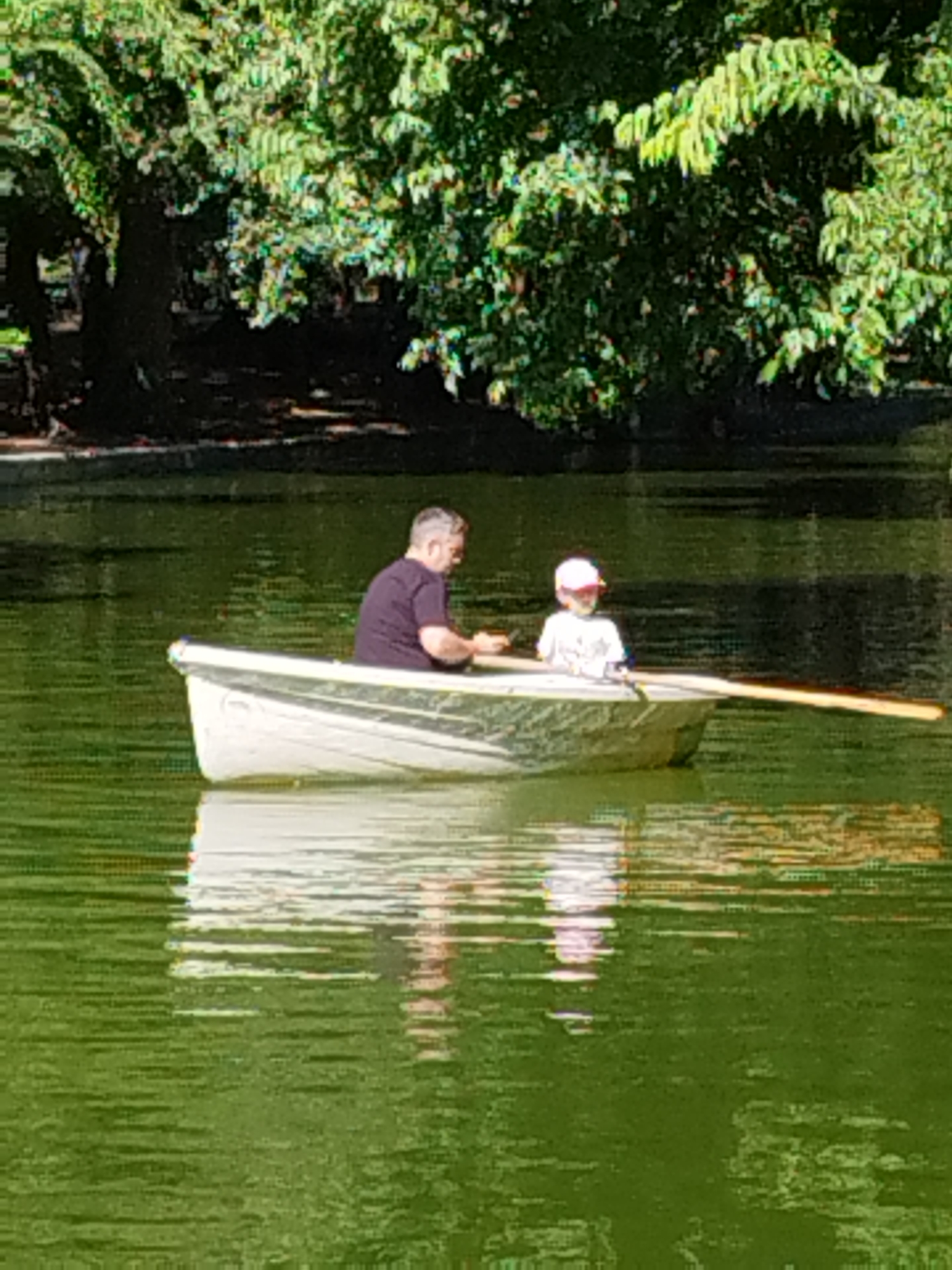 A man and a child are in a rowboat on a body of water. The man is in the rear of the boat, wearing a dark t-shirt and appears to be rowing. The child is in the front of the boat, wearing a light-colored t-shirt and a pink baseball cap. The boat is white with a green stripe along the side. The water is green and reflects the trees and sky. In the background, there are dense green trees.