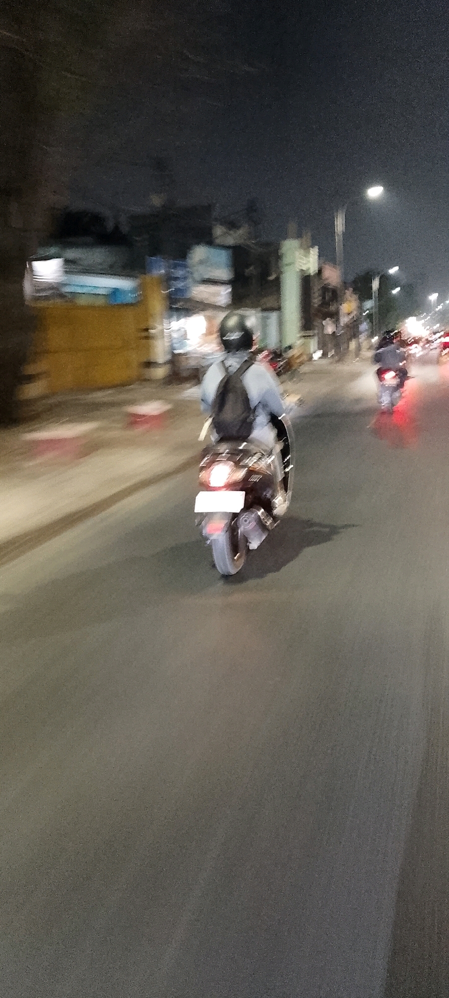The image, captured at night in Bekasi, Indonesia, shows a street scene with a person riding a motorcycle. The rider, wearing a dark helmet and a light-colored shirt, has a black backpack on. The motorcycle has its brake lights illuminated. The road is asphalt and appears wet or reflective, with a faint shadow cast by the motorcycle. In the background, there are buildings and streetlights, with other vehicles and lights visible further down the road, suggesting traffic. The overall image is motion-blurred, indicating the photograph was taken from a moving vehicle. The scene is illuminated by artificial light sources, typical of an urban environment at night.