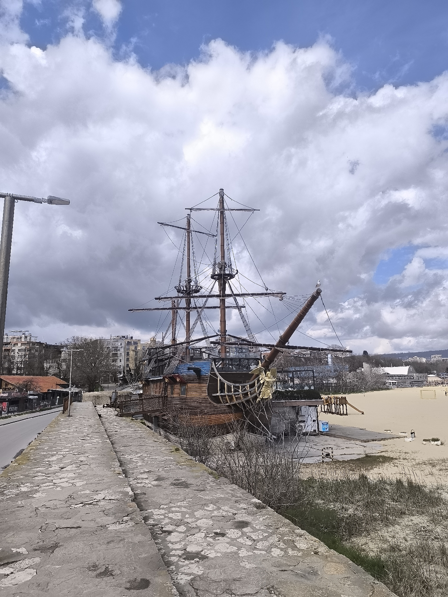 A daytime scene captures a large, non-sailing, wooden pirate ship structure, situated on a beachfront near a city. The ship, complete with masts and rigging, suggests a possible restaurant or tourist attraction. In the foreground, a stone walkway or barrier runs alongside a road and the beach, with some vegetation growing between the walkway and the sand. Buildings of Varna, Bulgaria are visible in the background behind the ship. The sky is overcast with dramatic cloud formations, suggesting a weather event. A street light post is on the far left. A single seagull is perched atop the bowsprit. A playground structure sits on the sand in the distance. No people are prominently featured.