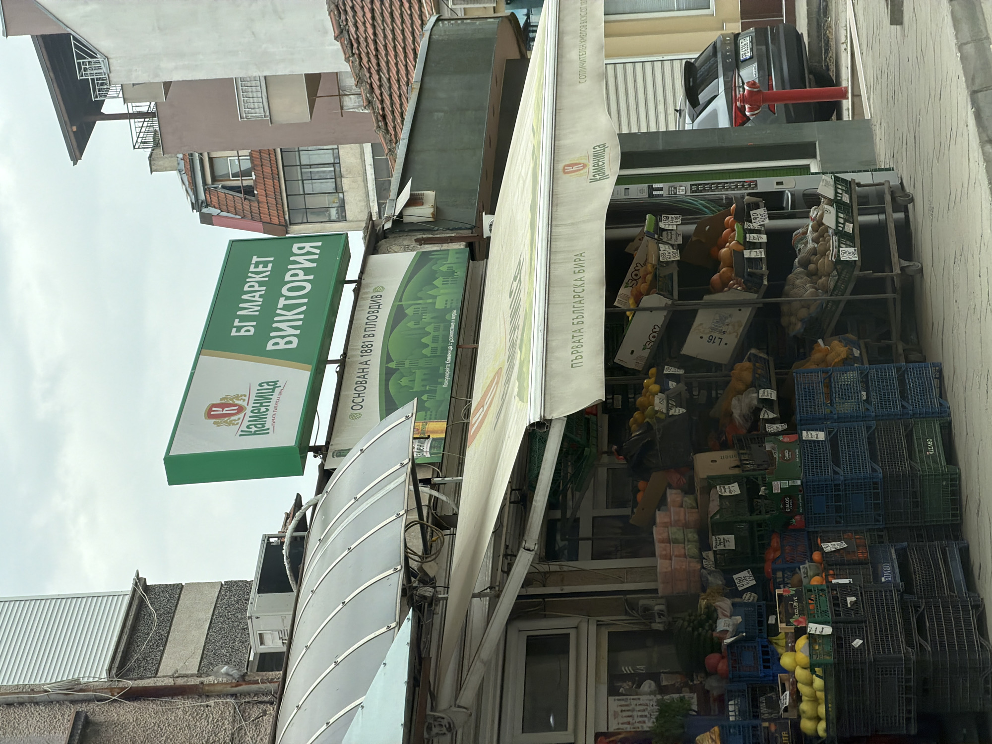 The image depicts a street scene, featuring a small outdoor market in Plovdiv, Bulgaria. The market is identified by a green sign reading "БГ МАРКЕТ ВИКТОРИЯ" (BG Market Victoria). Adjacent to this sign is another sign advertising "Каменица," a Bulgarian beer brand, with the slogan "ПЪРВАТА БЪЛГАРСКА БИРА" (The First Bulgarian Beer). The market stall displays a variety of fruits and vegetables in plastic crates and on metal shelves. Prices are marked on small signs. An awning with transparent panels covers the front of the market. Various buildings form the background, exhibiting a mix of architectural styles. A parked car is visible in the distance on the street.