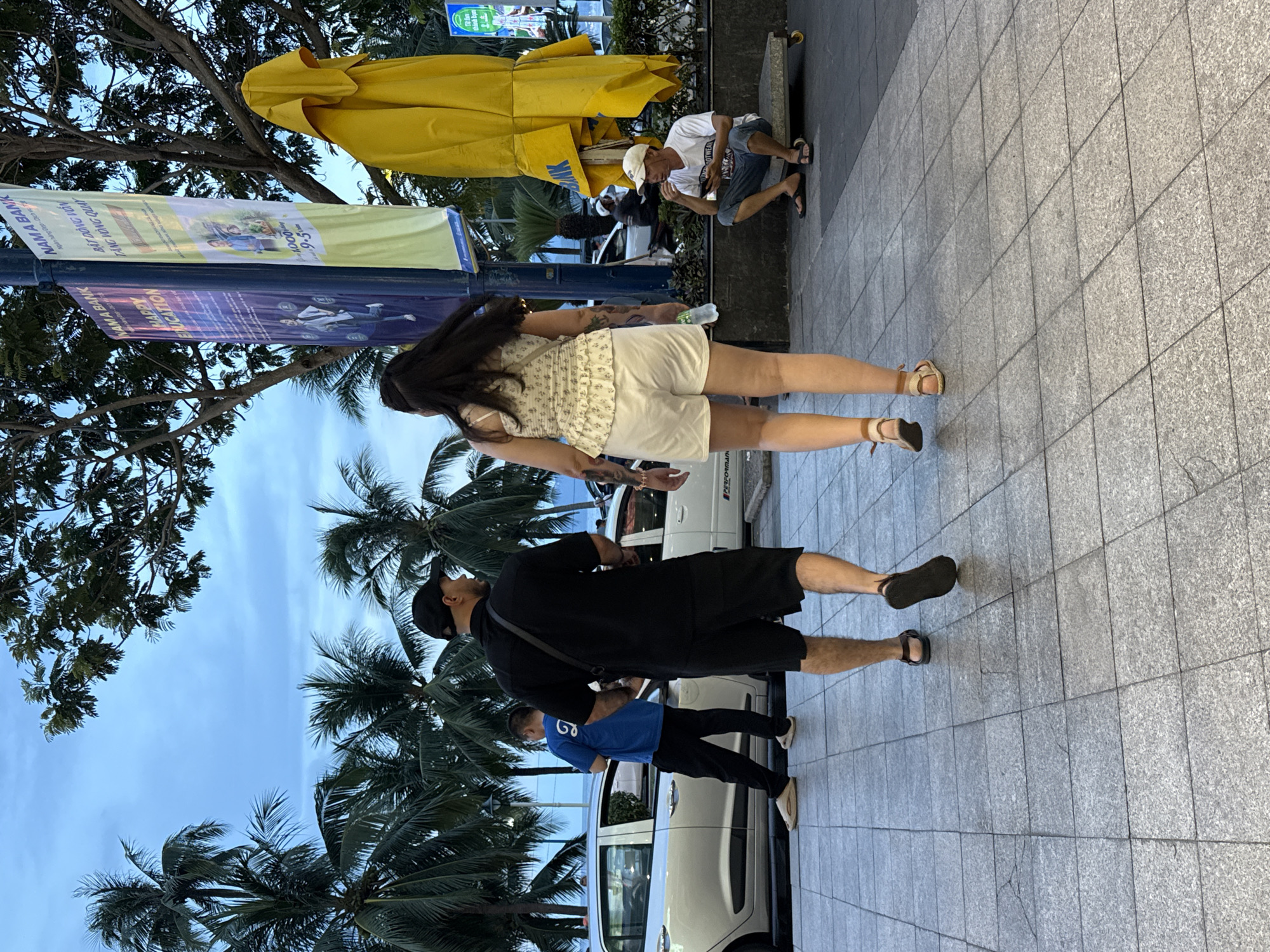 The image depicts a street scene in Nha Trang, Vietnam. The primary subjects are several people engaged in various activities along a sidewalk paved with granite tiles. In the foreground, a woman in a light-colored floral top and white shorts walks away from the camera, her legs and sandals clearly visible. Behind her, a man in a black t-shirt and shorts, also wearing sandals, is walking in the same direction. Further back, a person in a blue t-shirt and black pants is seen walking.
In the background, a man wearing a white t-shirt and shorts is sitting on a low bench, appearing to be eating. Yellow umbrellas, possibly part of a street vendor's setup or an outdoor dining area, are prominent in the upper left of the frame. Palm trees and lush green foliage are visible, suggesting a tropical or subtropical environment. The sky is overcast and appears to be either early morning or late afternoon.
Two banners are affixed to a lamppost. One banner, primarily yellow, displays text including "NAMA BANK" and "BẬT DÒNG VỐN TĂNG VÒNG QUAY" along with what appears to be business hours. The second, purple banner, also features "NAMA BANK" and the text "HAPPY EDUCATION." The presence of cars in the background, including a white vehicle, indicates a street or public area. The overall scene suggests a casual, everyday moment in a bustling urban environment.