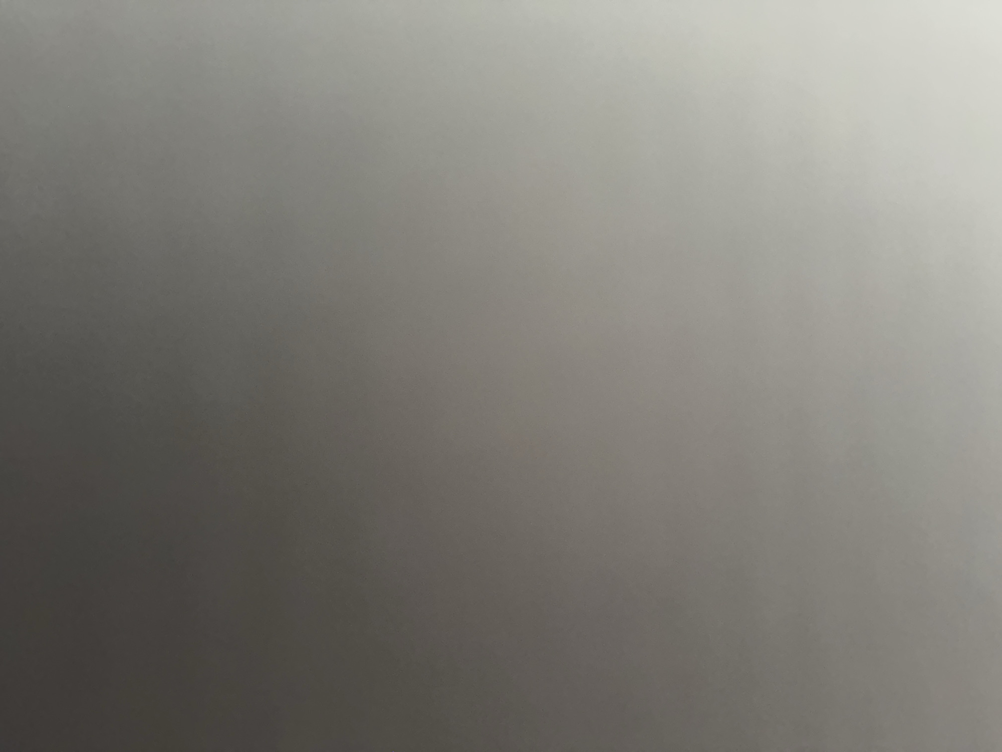 This is a still photograph, likely taken indoors, of a plain, light grey surface. The lighting in the image creates a subtle gradient, with the left side appearing darker and the right side becoming progressively lighter and brighter. There are no discernible subjects, objects, or activities taking place. The texture of the surface appears smooth, with a slight grain visible upon closer inspection, suggesting it could be a wall, a piece of fabric, or a similar material. There is no visible text, indication of time of day, weather, or specific location cues beyond the general context of Belgrade, Serbia. The overall impression is one of a simple, uncluttered background.