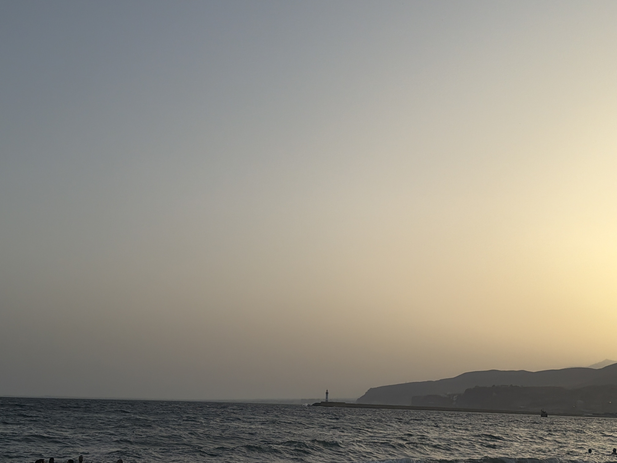 A wide shot of a coastline at dusk features a calm sea with gentle waves in the foreground. A distant lighthouse stands on a pier extending into the water, with hazy hills forming the background. The sky transitions from a pale blue at the top to a warm yellow near the horizon. A few people are visible near the water's edge in the bottom left, and a small boat can be seen in the distance on the right.