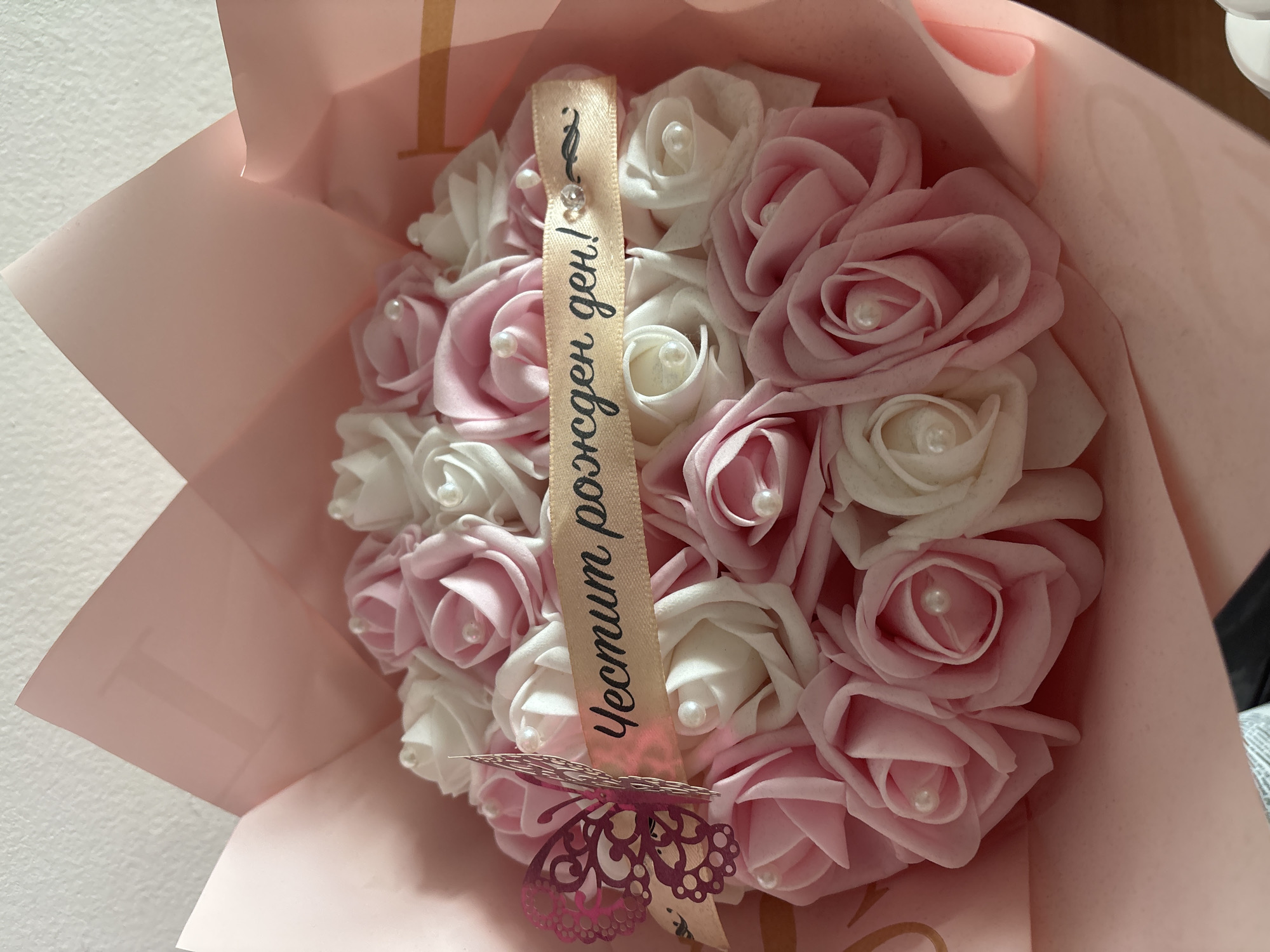 A close-up shot of a bouquet of artificial roses. The roses are arranged in a dome shape, with alternating colors of pink and white. Each rose has a small pearl in the center. The bouquet is wrapped in light pink paper. A ribbon with Cyrillic script on it is draped over the roses. A butterfly-shaped decoration is pinned to the ribbon. The background is a plain white wall. The bouquet appears to be located indoors. There is no indication of Troyan, Bulgaria in the image.
