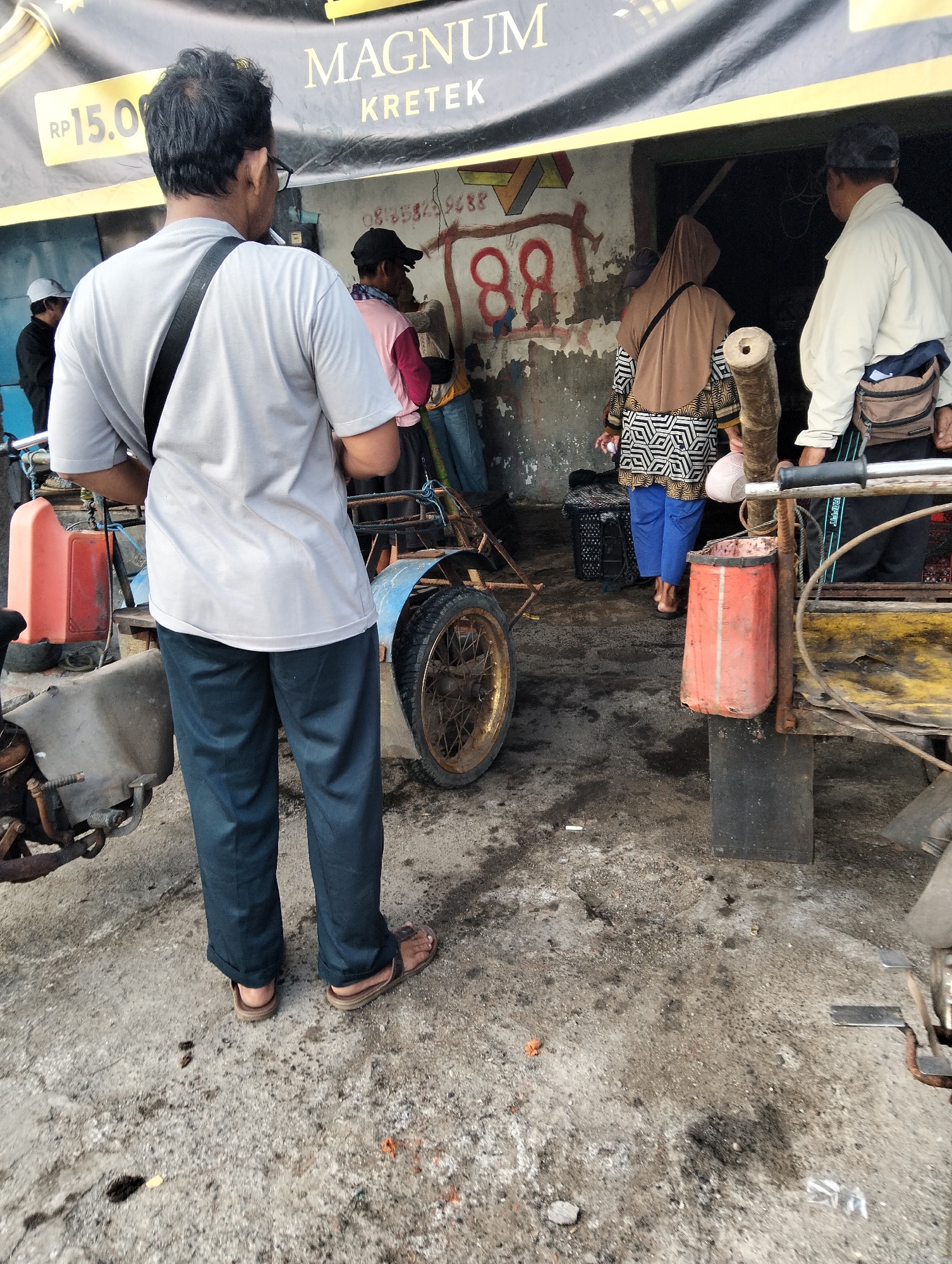 The image depicts an outdoor scene in Muncar, Indonesia, likely in front of a shop or stall. In the foreground, a man wearing a grey t-shirt and dark blue pants stands with his back to the camera, facing a rustic structure with a banner overhead. The banner displays the word "MAGNUM KRETEK" in large lettering, with "RP 15.000" visible on a yellow price tag on the left side. Behind him, other individuals are visible. A person in a pink shirt and black cap is partially obscured, and a woman in a brown hijab and a patterned dress is seen walking away. Another person, wearing a light-colored jacket and a cap, stands to the right, facing into the structure.
The environment appears somewhat unkempt, with a concrete ground that is stained and littered with debris. Several wheeled carts or vehicles, likely used for transport or selling goods, are present. One cart in the foreground has a rusty metal body and a large spoked wheel. Another cart to the left has a bright orange cylindrical object attached to it, possibly a container or fuel tank. The structure behind the people appears to be made of rough materials, with peeling paint and graffiti on the wall. The overall impression is of a busy, informal commercial area.