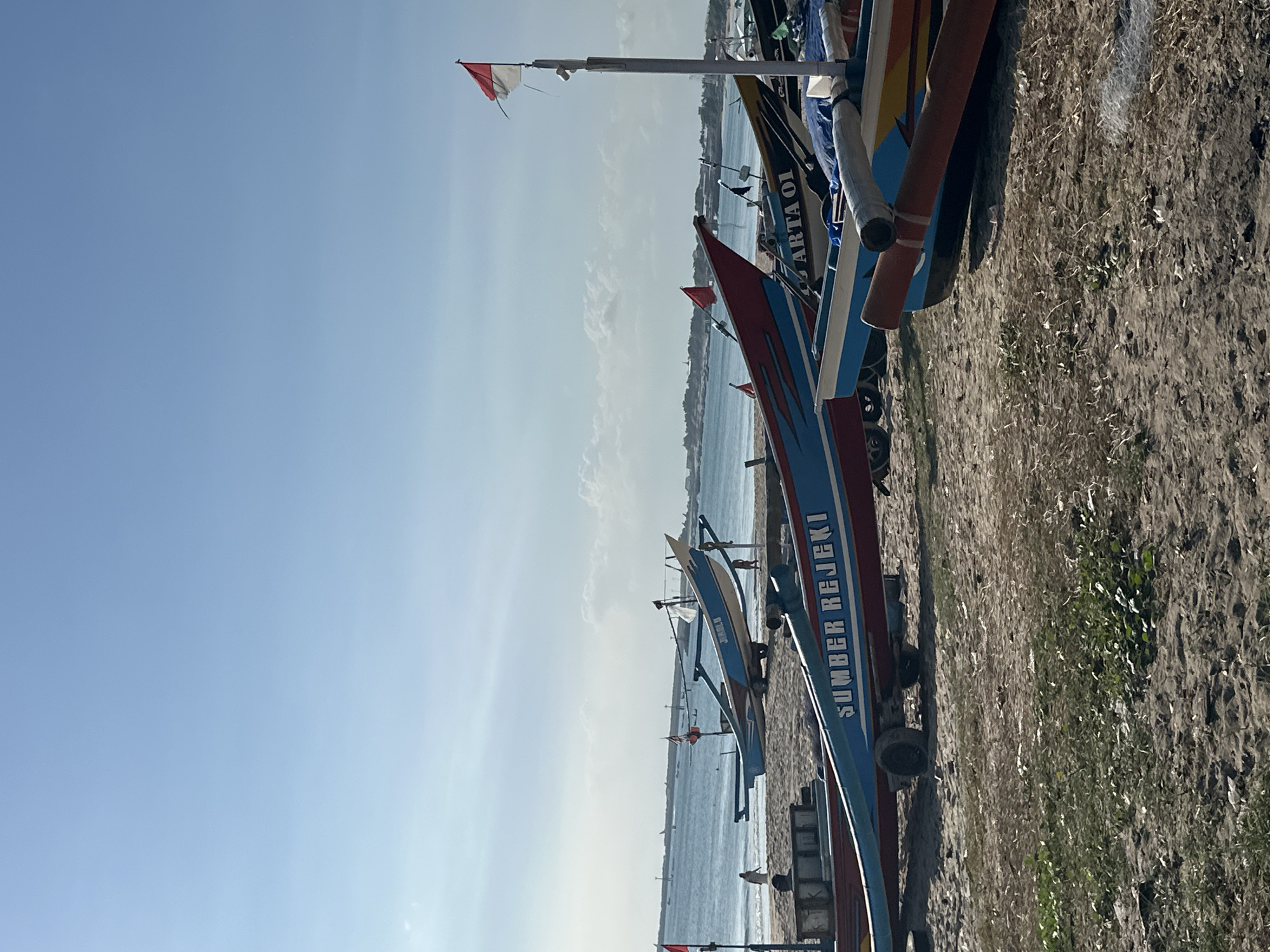 A group of traditional Indonesian fishing boats are beached on a sandy shore in Kuta, Indonesia, with the ocean visible in the background under a clear blue sky. The boats are colorful, with several displaying writing on their sides, such as "SUMBER REJEKI" and "ARTA 01". One boat prominently features the Indonesian flag. The shore is covered in sand, with some patches of sparse vegetation. There are a few people visible walking on the beach. The overall scene suggests a typical day for these fishing vessels.