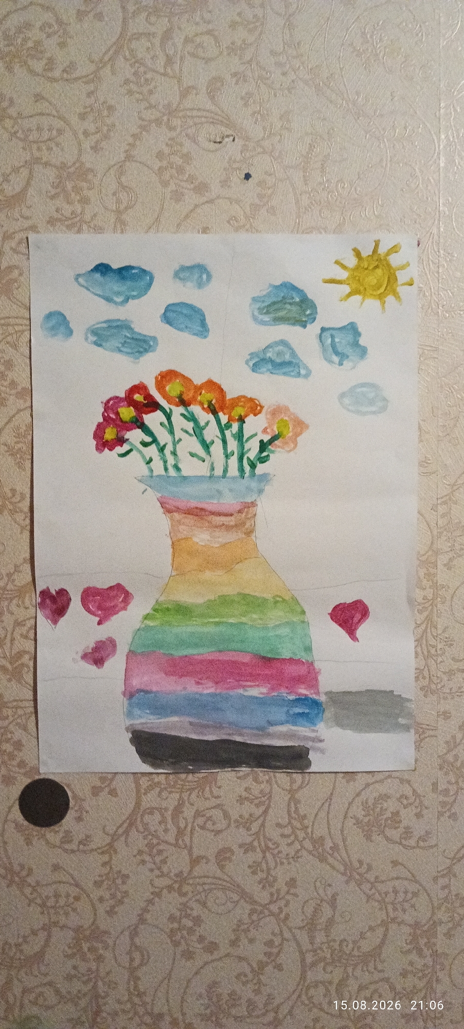 The image shows a watercolor painting of a vase with flowers, clouds, and a sun. The vase is depicted with horizontal stripes of various colors, including yellow, orange, pink, green, blue, and black. The flowers are a mix of red, pink, and orange, with green stems. The sky is filled with blue clouds and a bright yellow sun. There are also three pink hearts on the left side of the painting and one pink heart on the right. The painting is on a piece of white paper, which is placed against a wall with a patterned wallpaper. A date and time stamp, "15.08.2026 21:06", is visible in the bottom right corner. The scene appears to be an indoor setting, with the painting propped up on a wall. There is no indication of the city of Mahilyow, Belarus in the image itself.